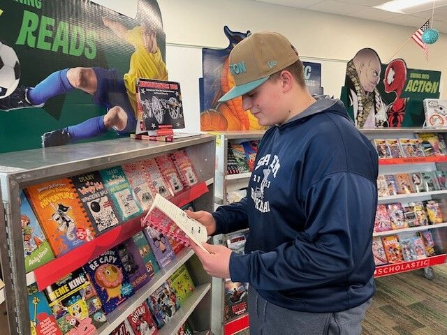 A young person wearing a baseball cap and hoodie browses books at a Scholastic book fair.