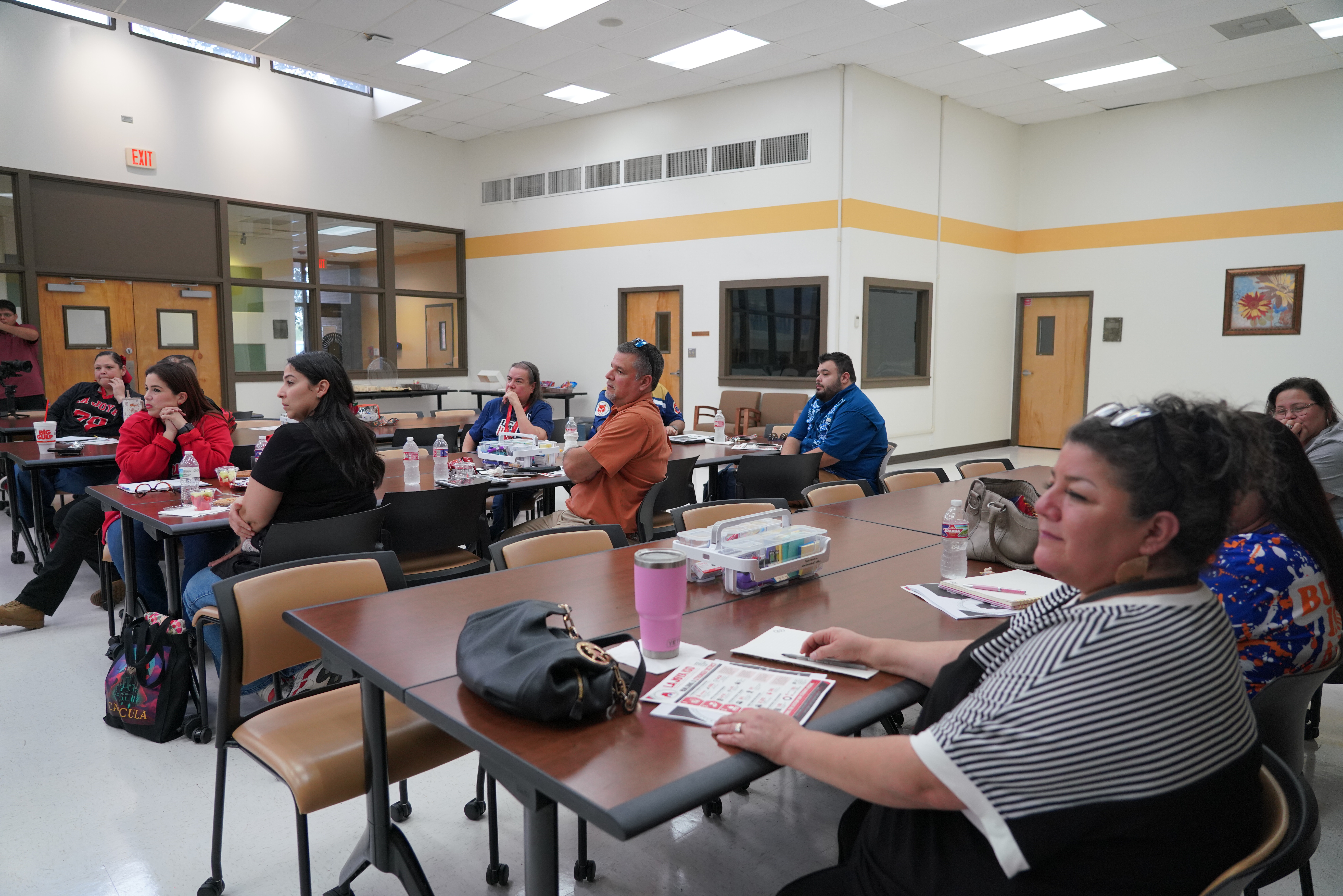A group of adults sits at tables in a bright room, attending a meeting or presentation.
