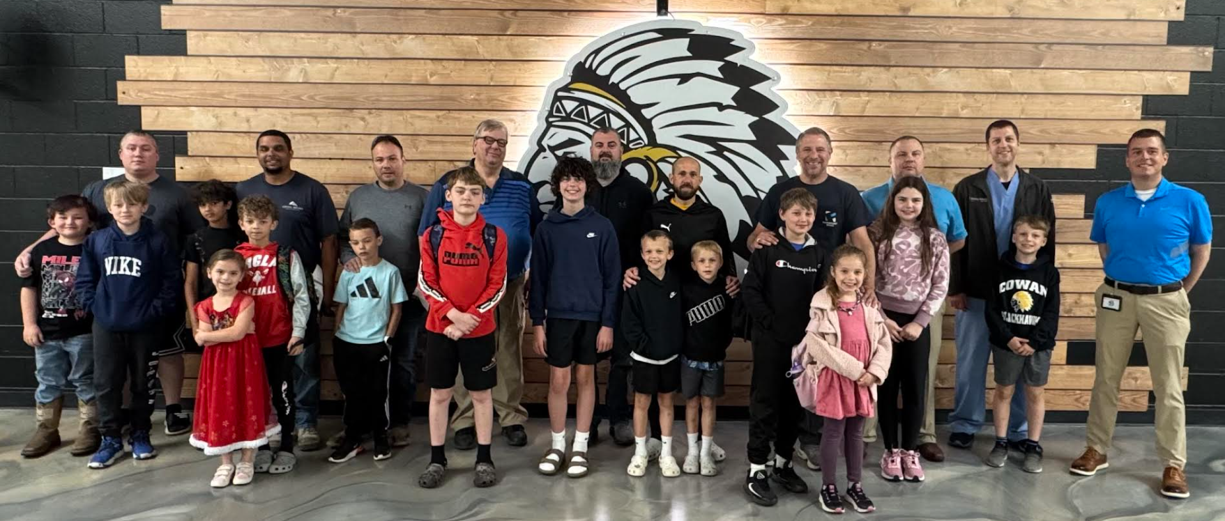 A group of adults and children pose for a photo in front of a wood-paneled wall with a Native American-inspired graphic.