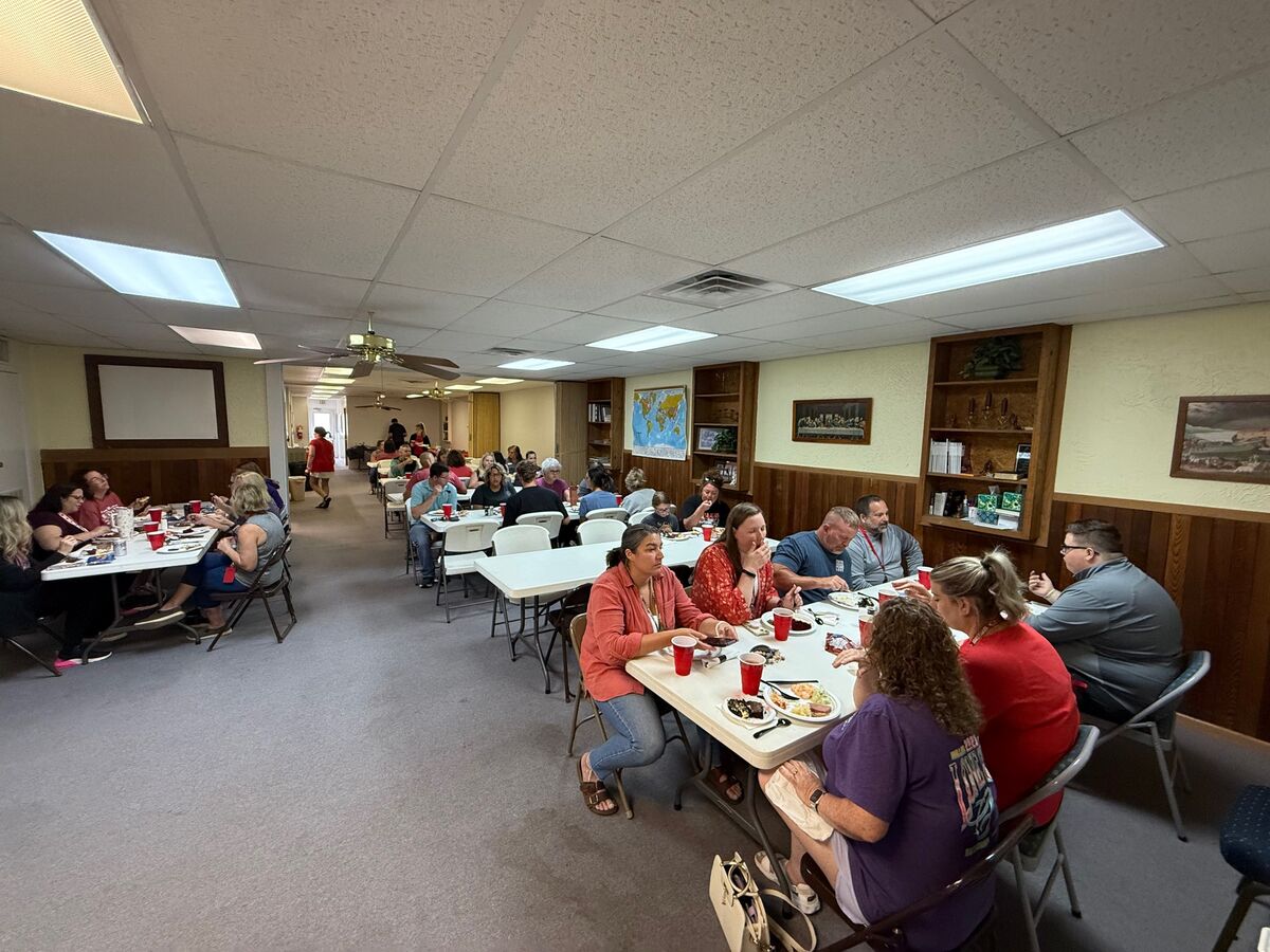 A group of people are seated at long tables, enjoying a meal together in a room with fluorescent lighting.