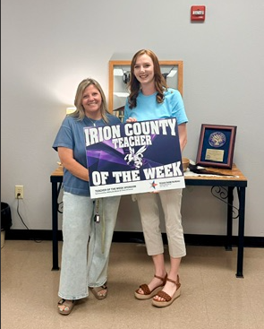 Two women stand together holding a sign that reads 'IRION COUNTY TEACHER OF THE WEEK'.