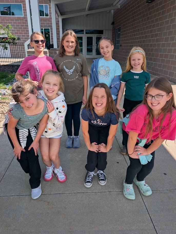 A group of eight young girls smile happily together on a sunny outdoor sidewalk.