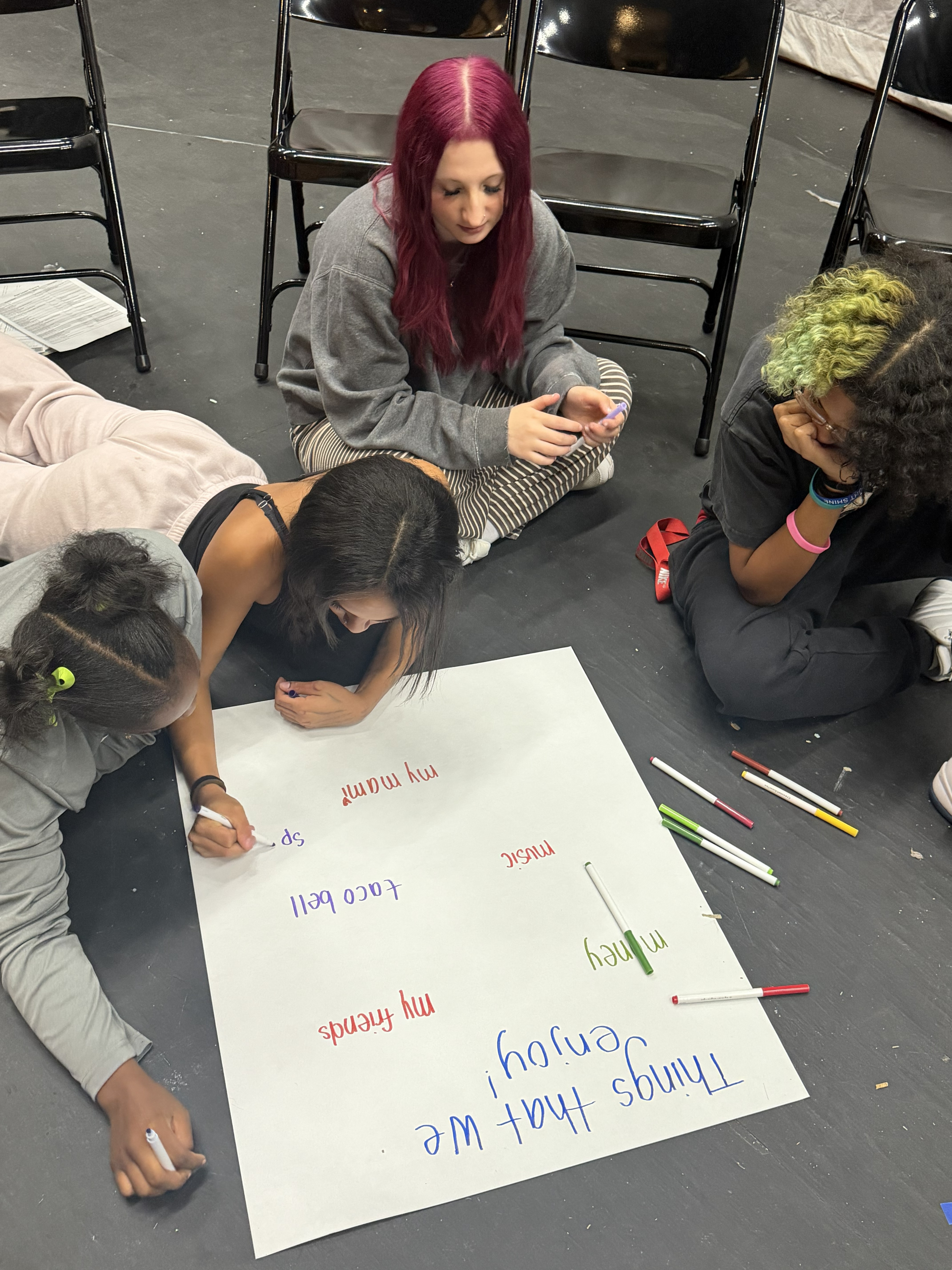 Four young people collaborate on a large poster on the floor, writing with colorful markers.