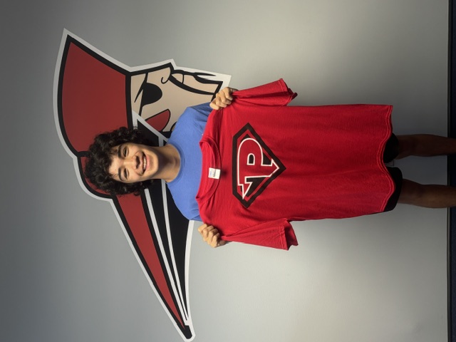 A smiling young person holds up a red t-shirt with a large letter P logo, standing in front of a wall with a graphic decal.
