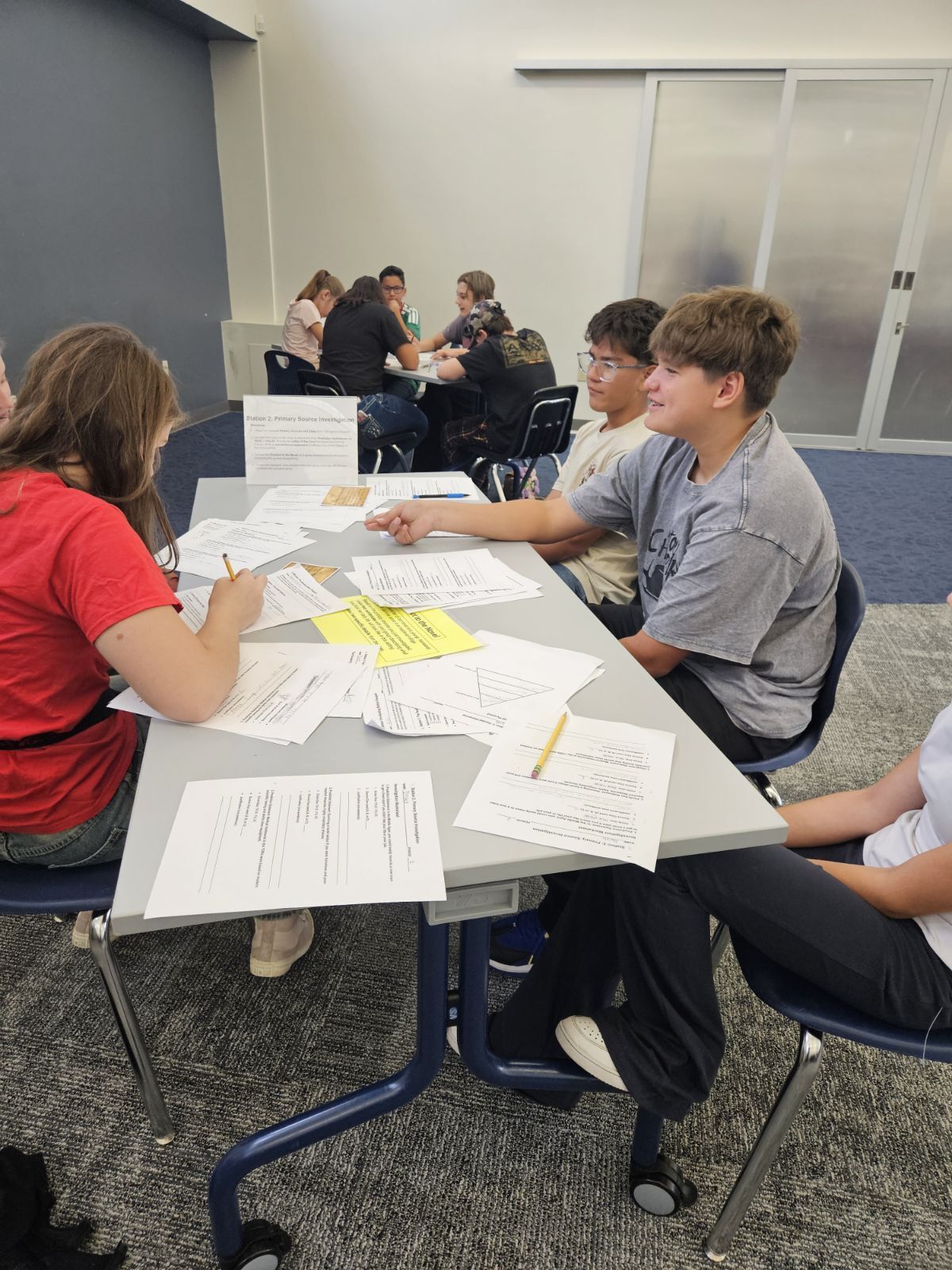 Students collaborate around a table filled with papers and worksheets during a class activity.