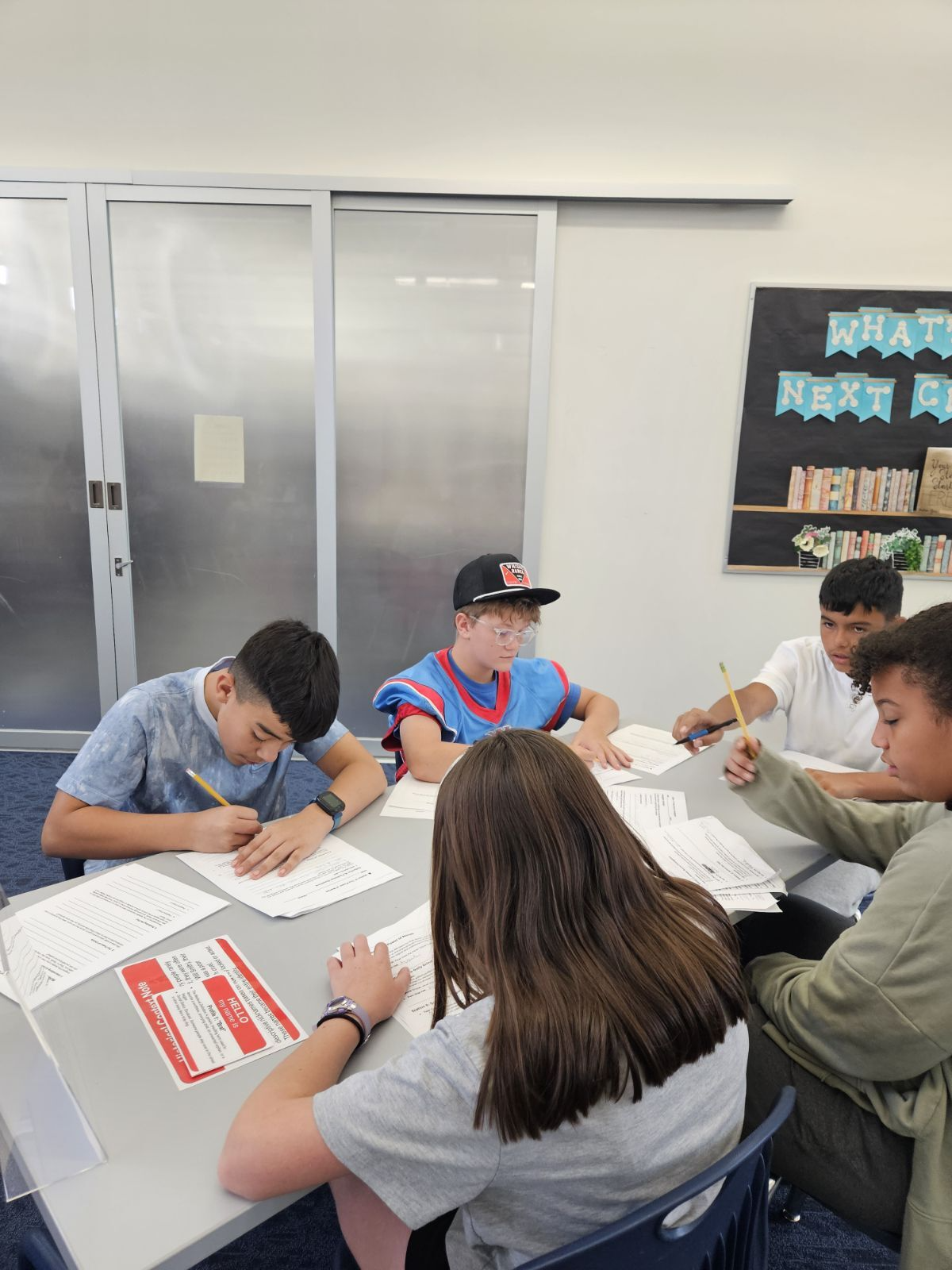 Students work together at a table, writing on papers with pencils.