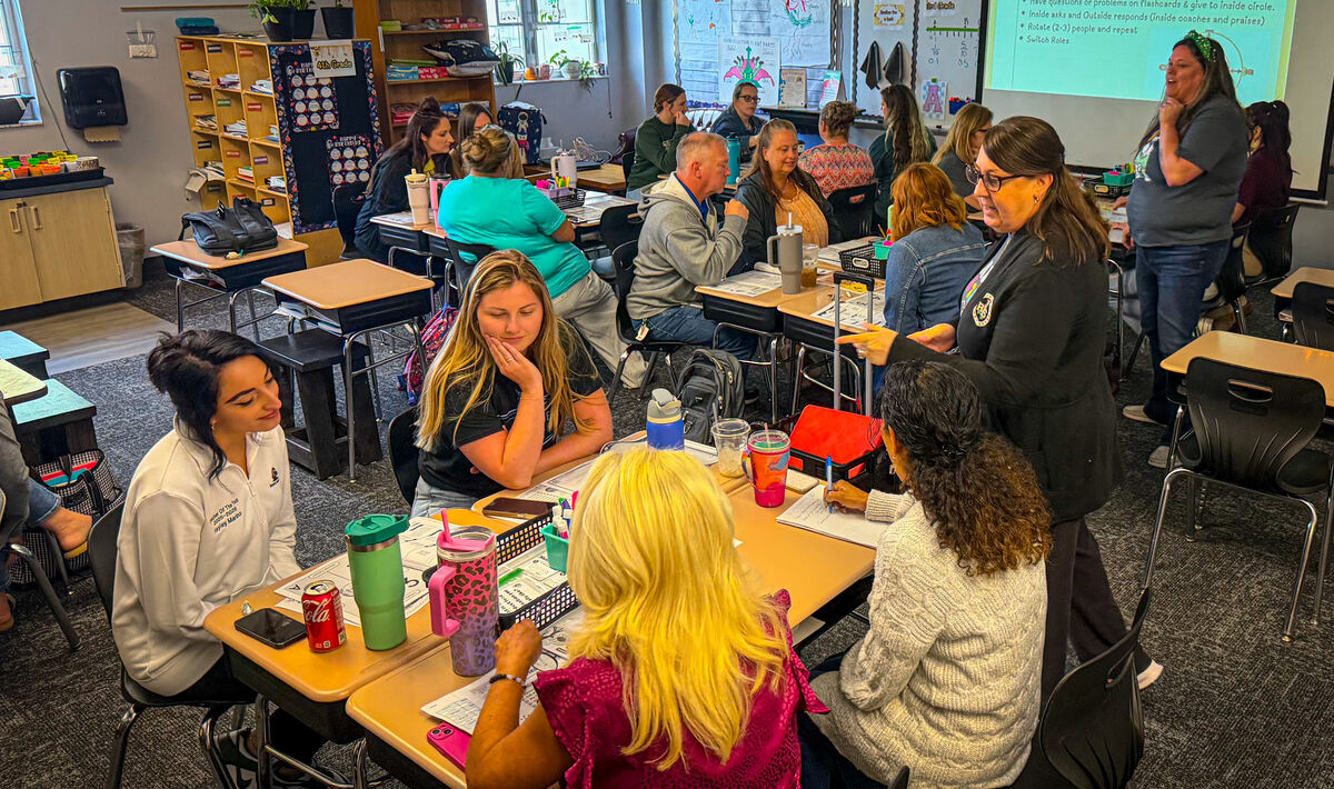 A group of adults are gathered in a classroom, seated at desks and listening to a speaker.