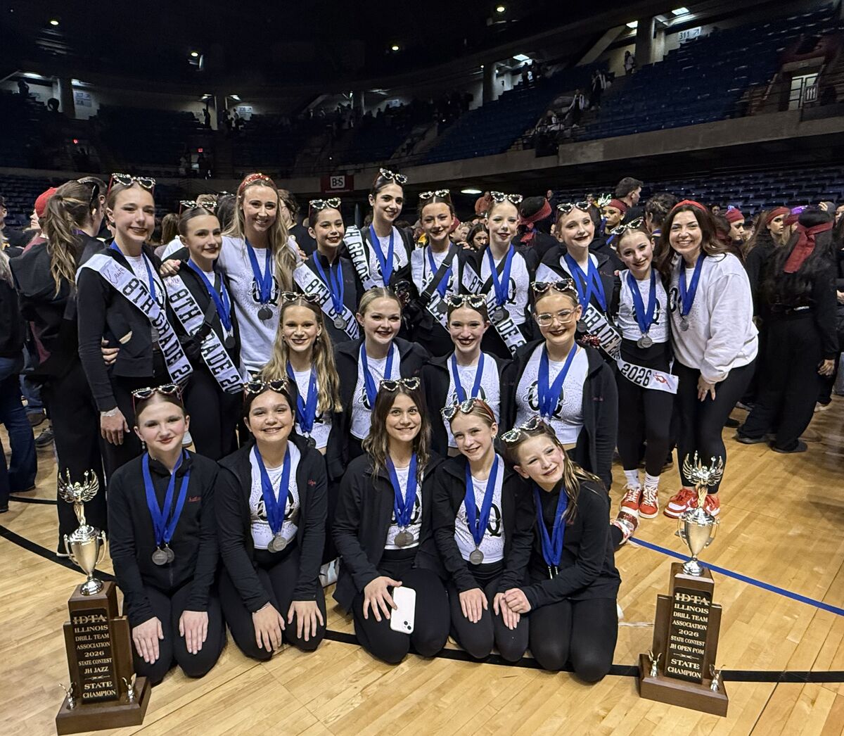 A group of young dancers in matching black outfits and sparkly sunglasses pose with trophies and medals.