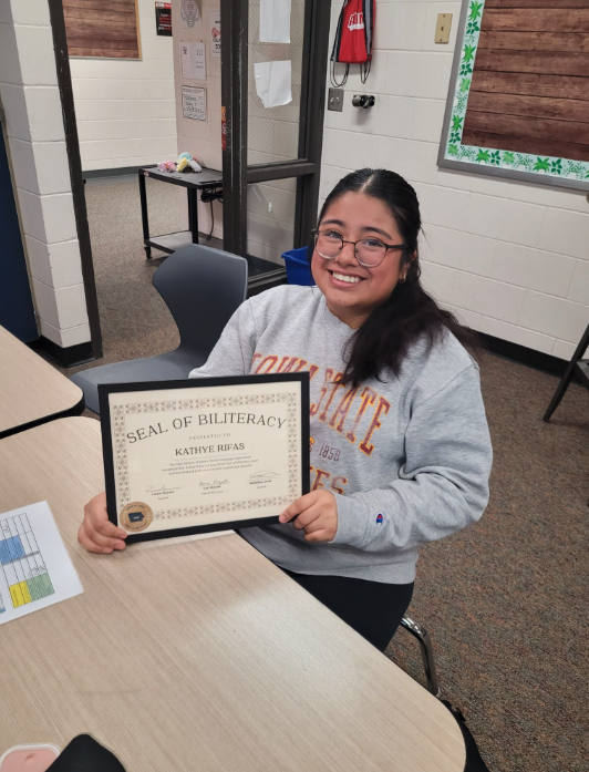 A smiling young woman proudly holds a framed Seal of Biliteracy certificate.