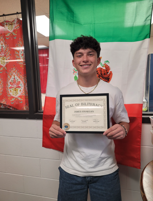 A young man smiles proudly while holding a Seal of Biliteracy certificate in front of a green, white, and red striped flag.