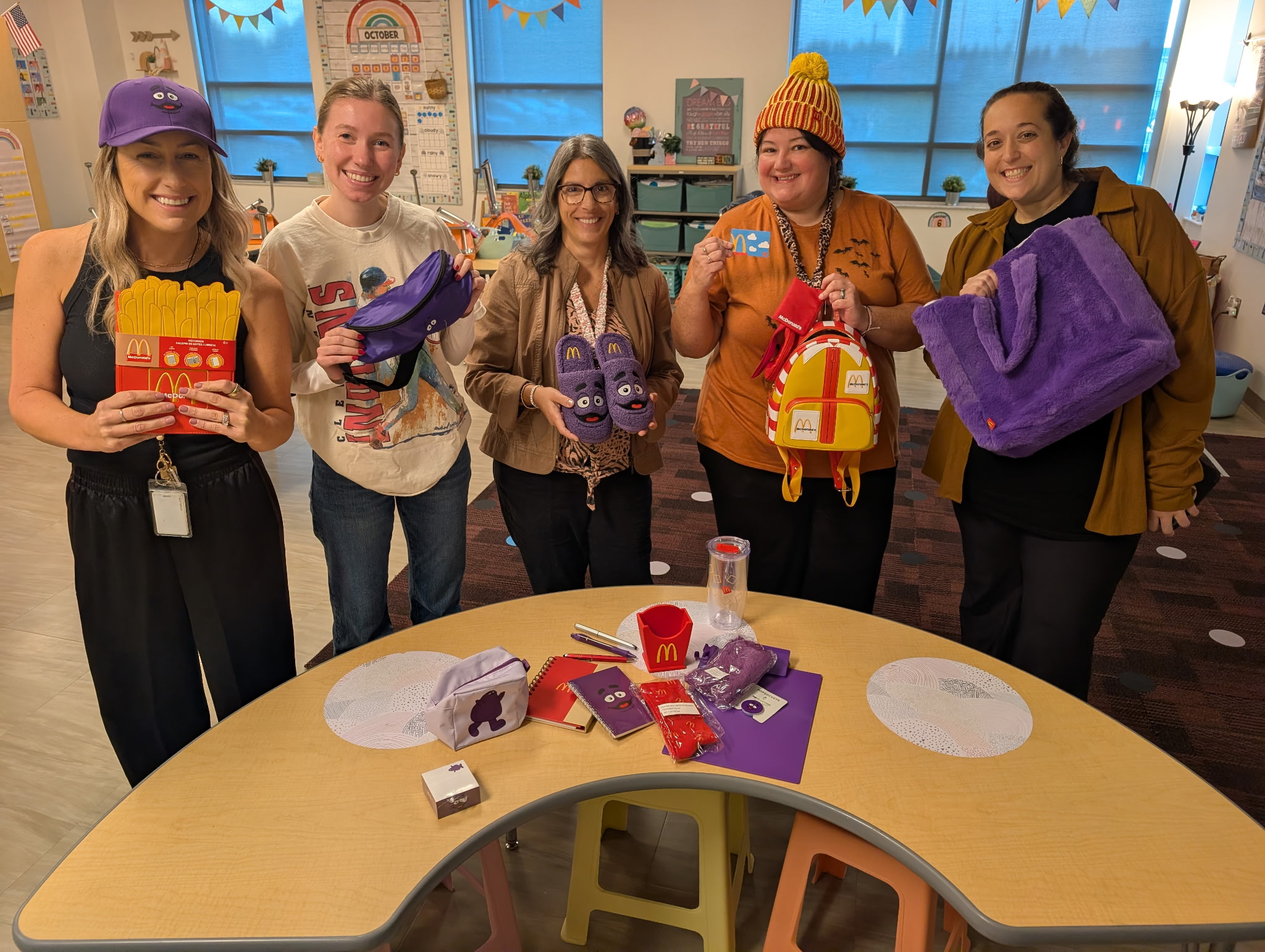 Five adults stand smiling in a classroom, holding McDonald's Grimace-themed merchandise around a curved table with more items.