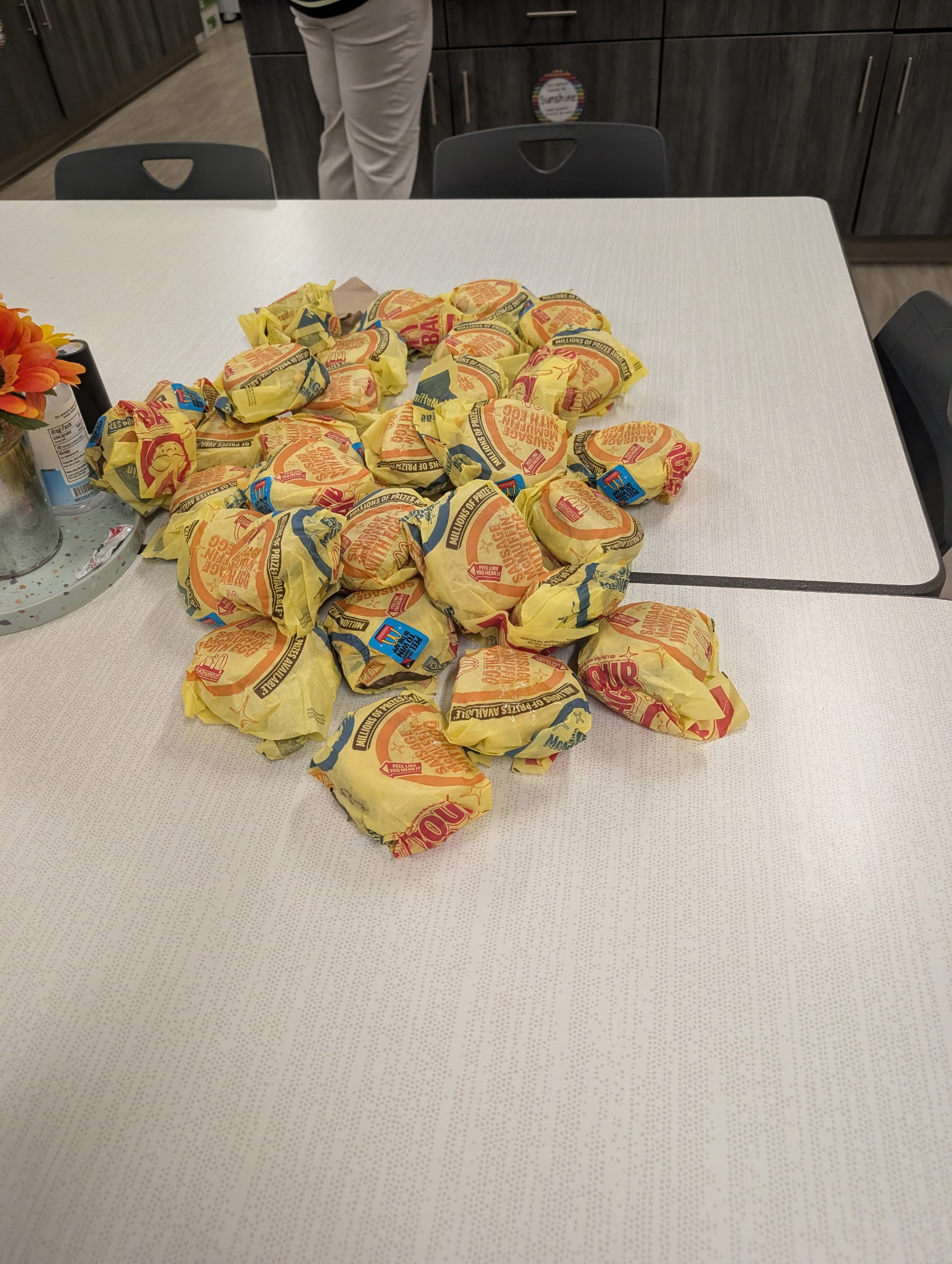 A large pile of wrapped breakfast sandwiches rests on a white table in a breakroom.