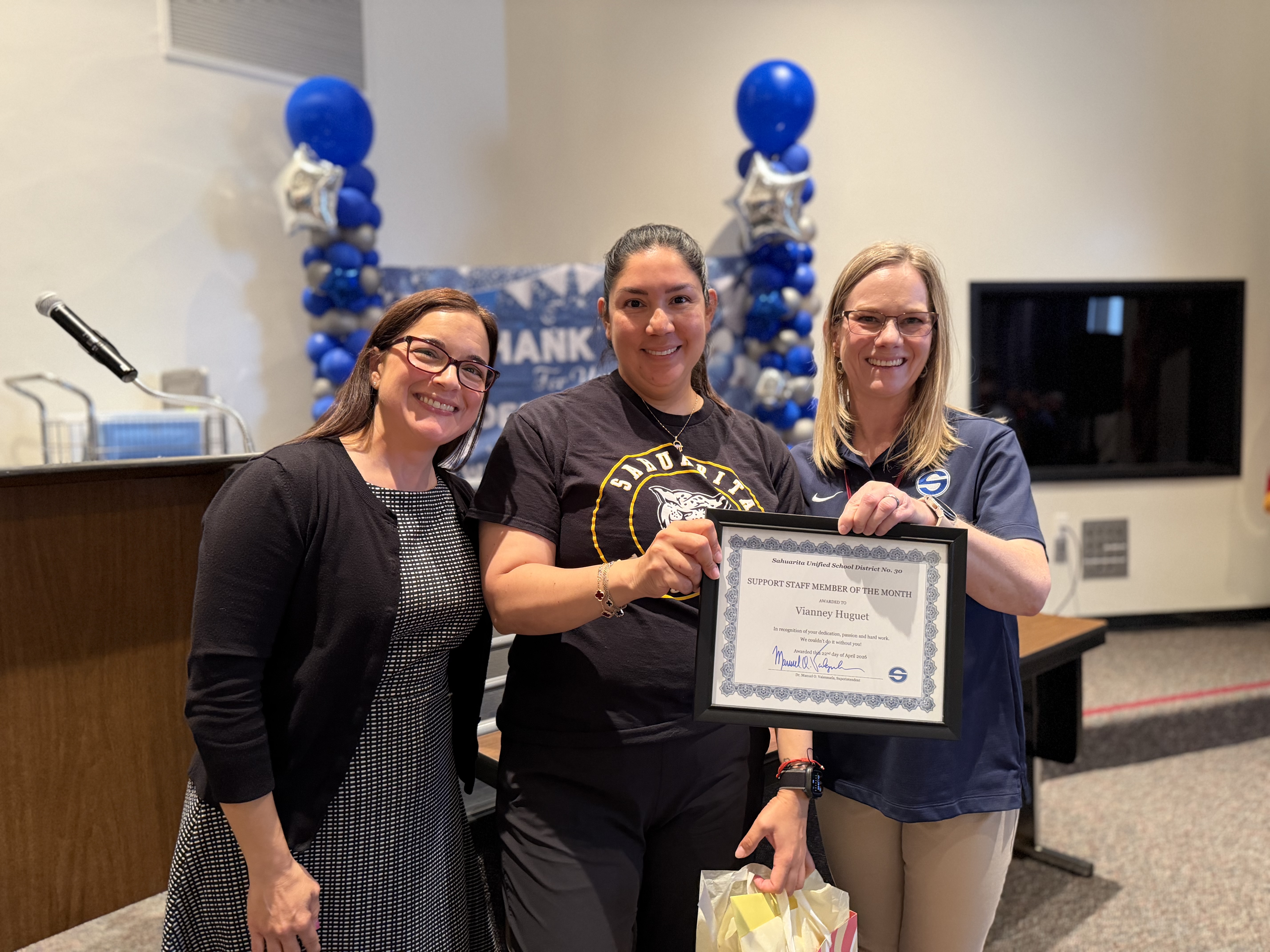 Three women smile as one holds a framed certificate for "Support Staff Member of the Month" award
