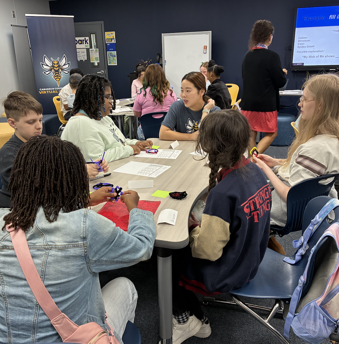 Students work on a craft project at a table, with a teacher presenting at the front of the room.