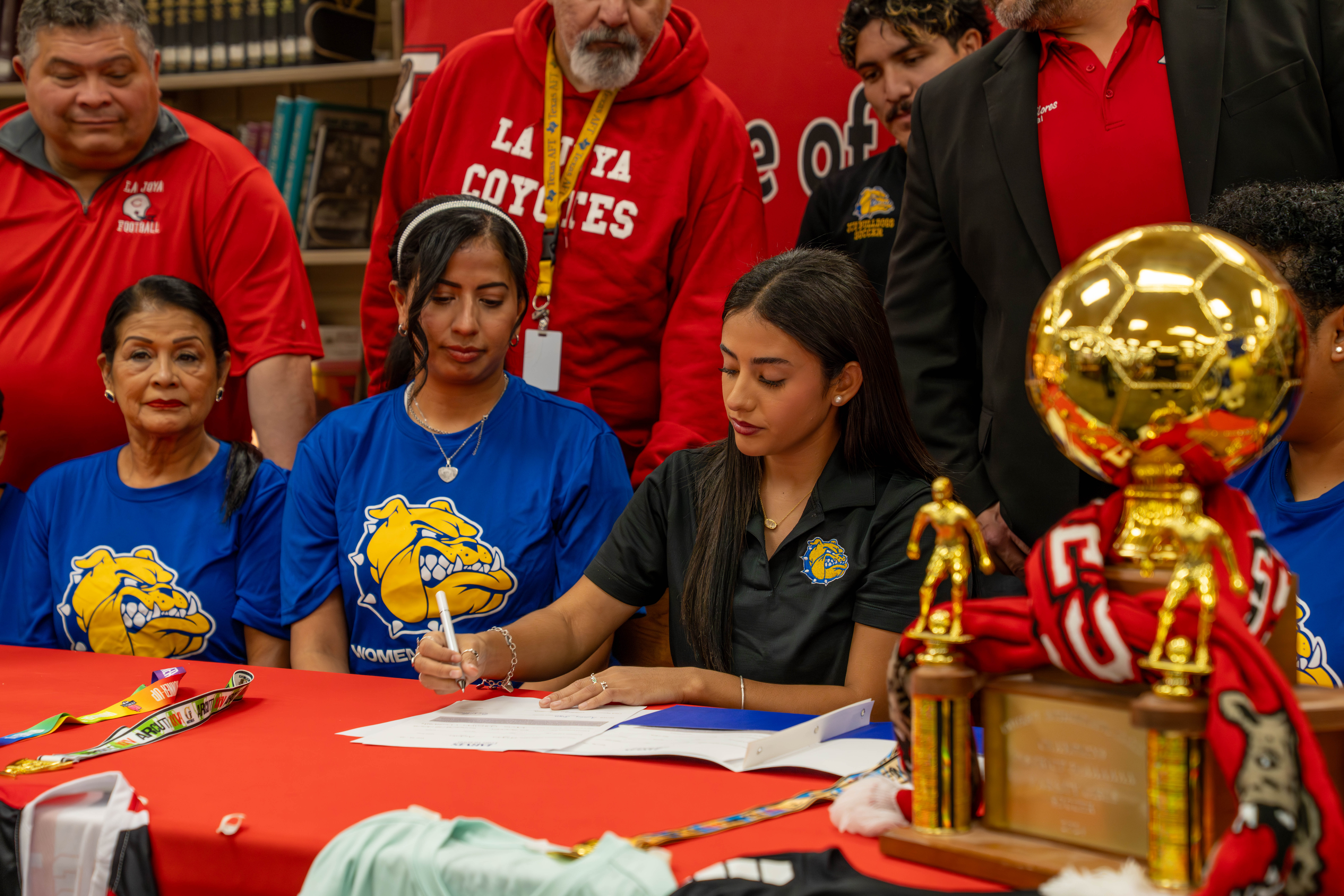 A young woman signs a document at a red table, surrounded by supporters and athletic trophies.