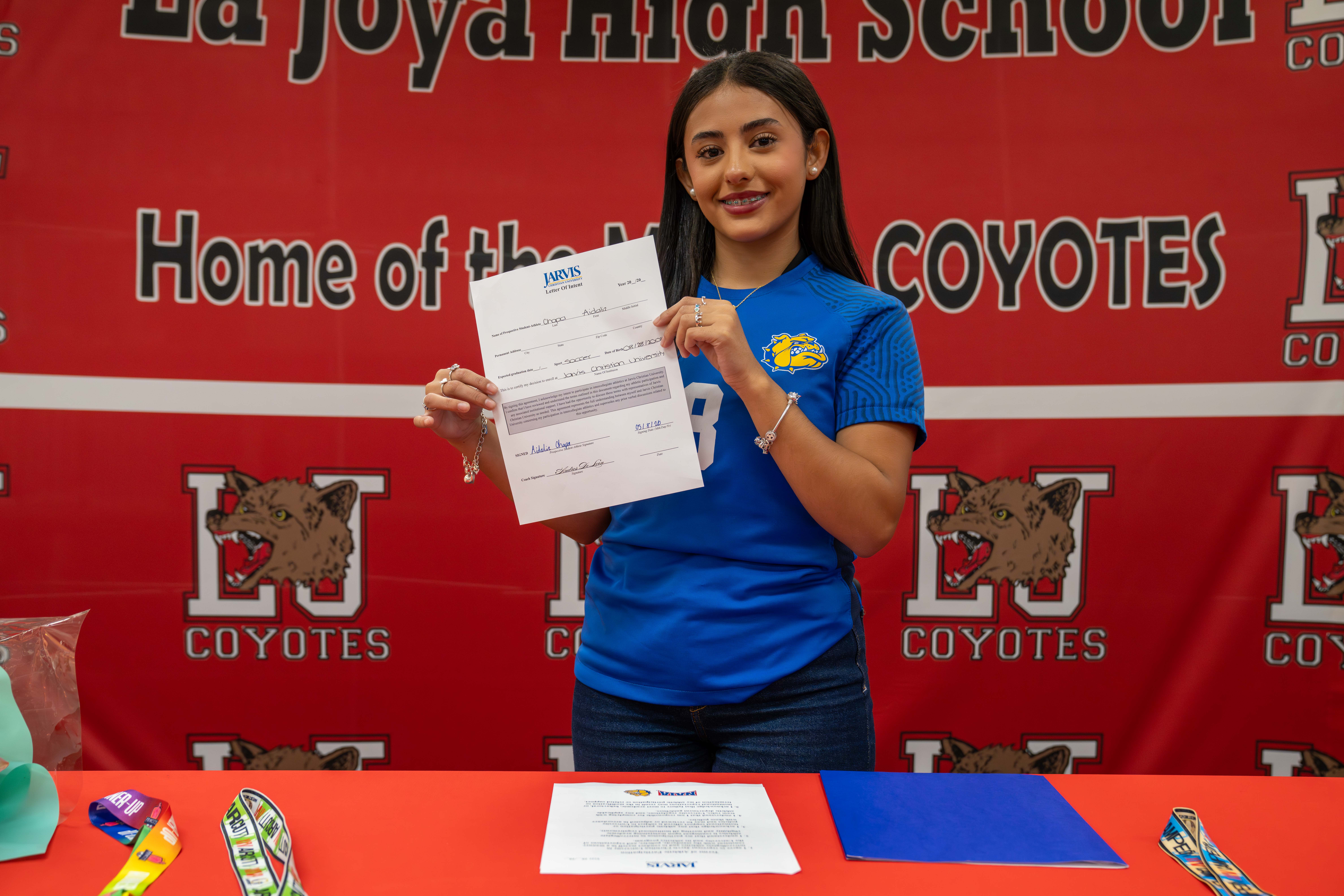 A young woman in a blue shirt smiles while holding a "Letter of Intent" document in front of a red banner with "La Joya High School Coyotes" text.