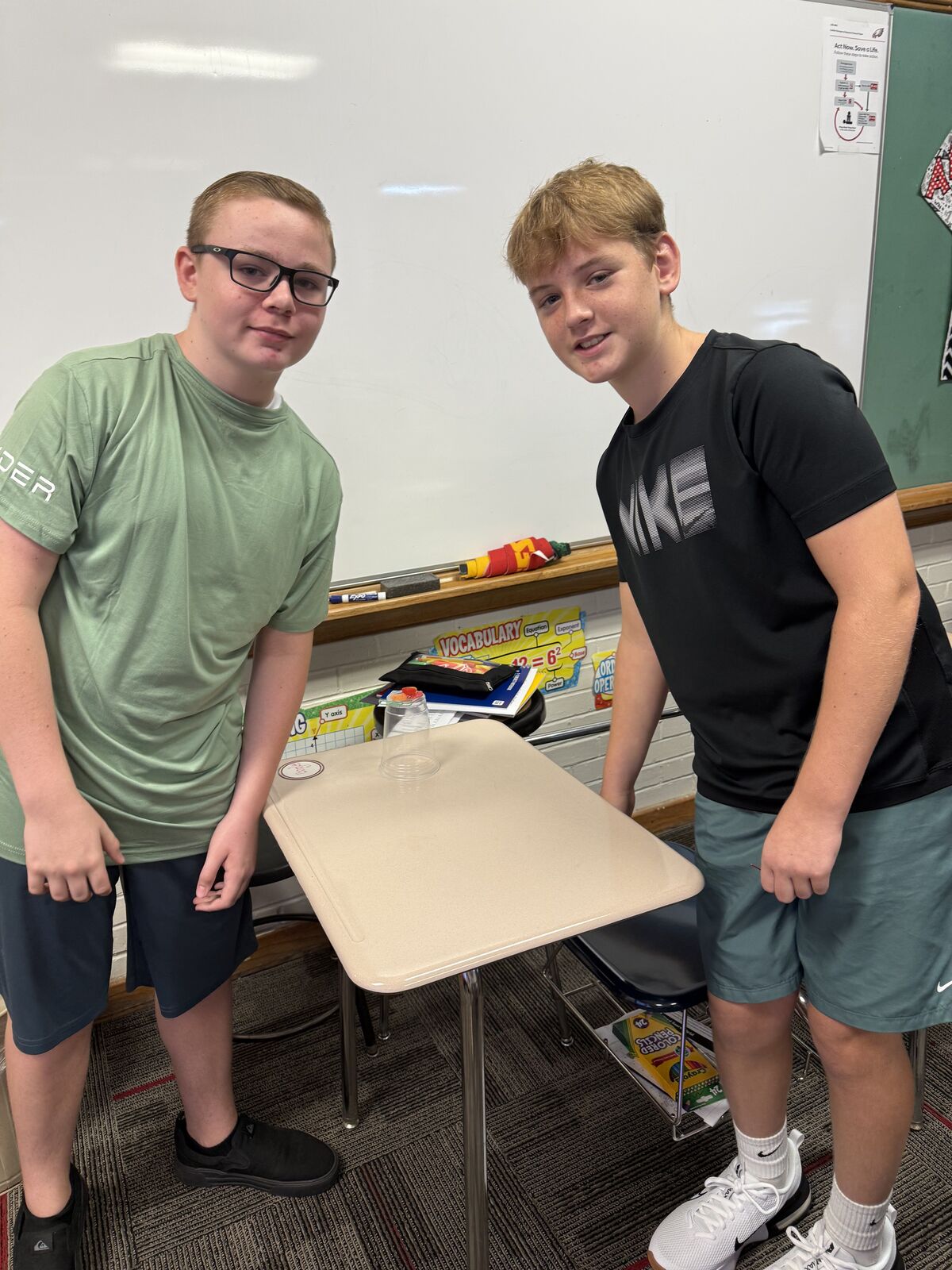Two young people stand near a whiteboard in a classroom setting.
