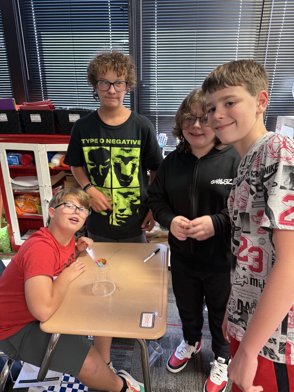 Four young students are gathered around a desk in a classroom, looking at the camera.