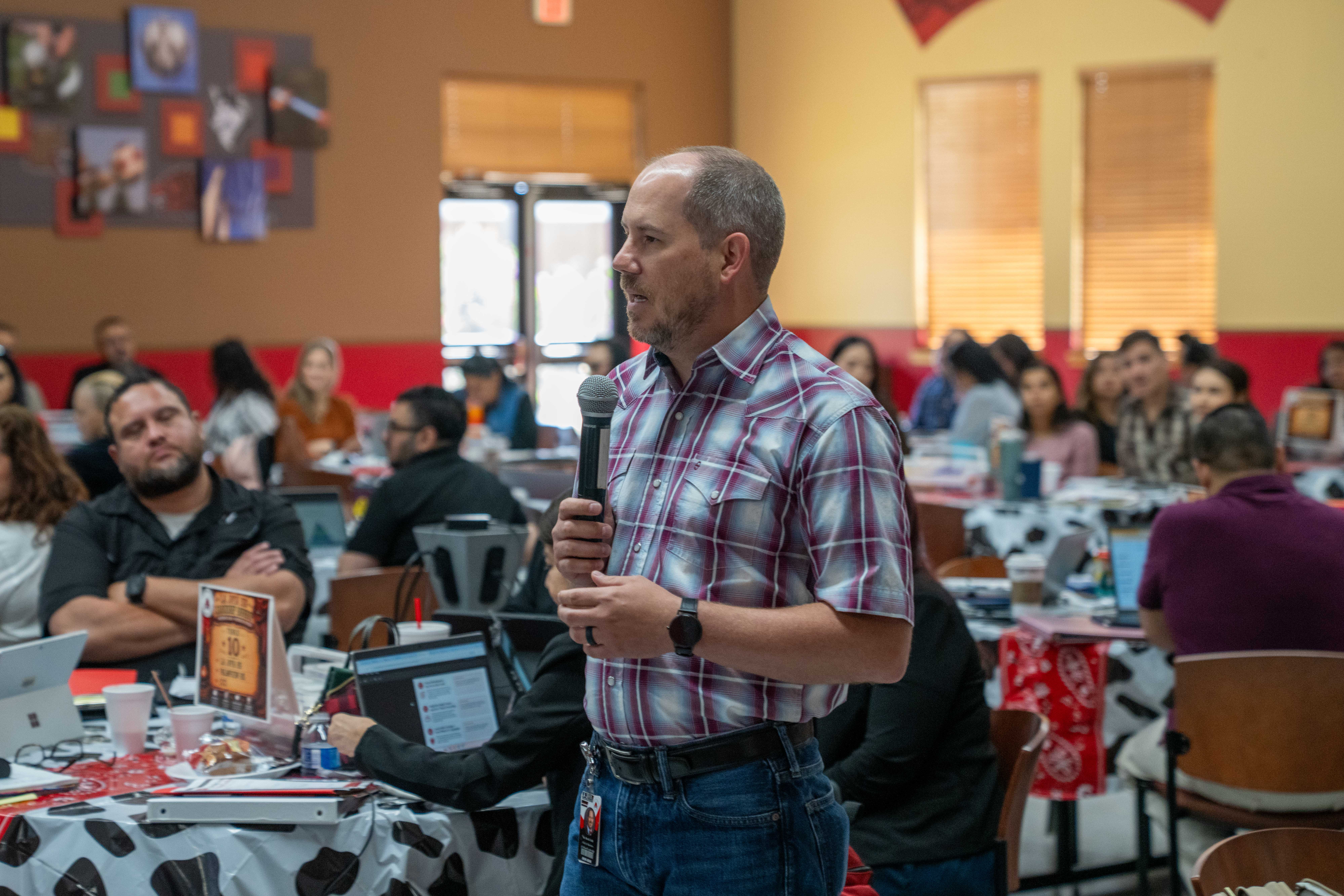 A man in a plaid shirt speaks into a microphone while addressing an audience seated at tables.