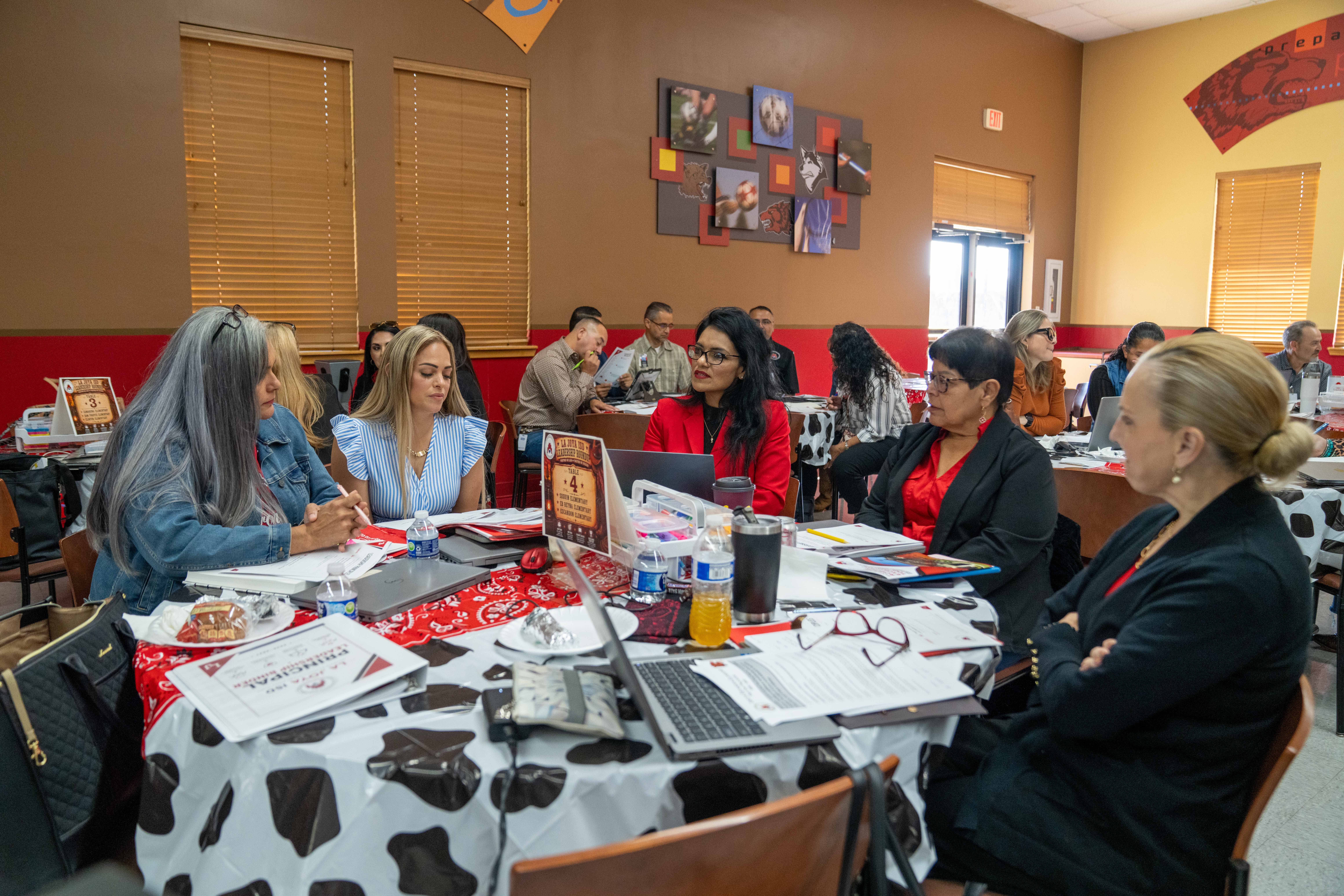 A group of people sit around a table covered with a cow-print tablecloth, engaged in a meeting with laptops and papers.