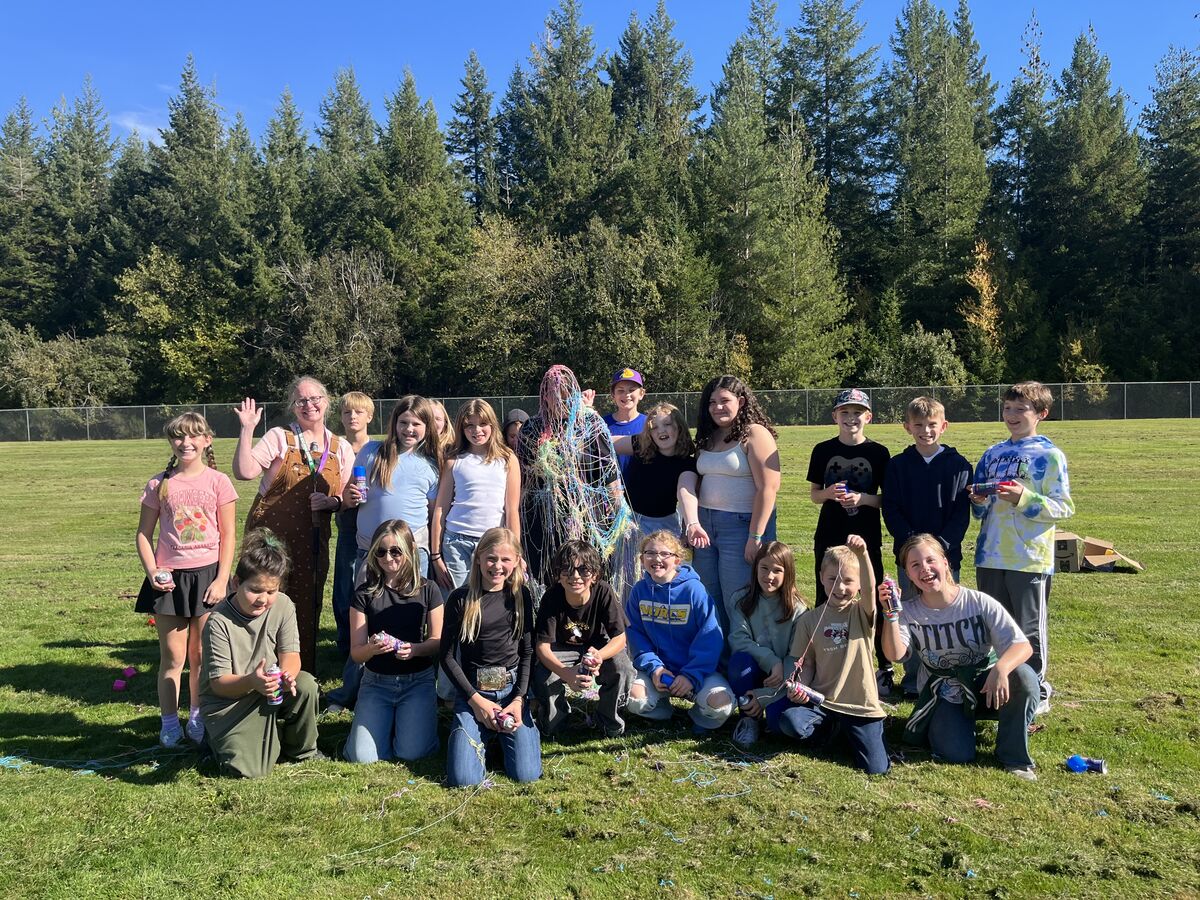 A group of children and adults pose on a sunny grassy field, with one person playfully covered in colorful silly string.