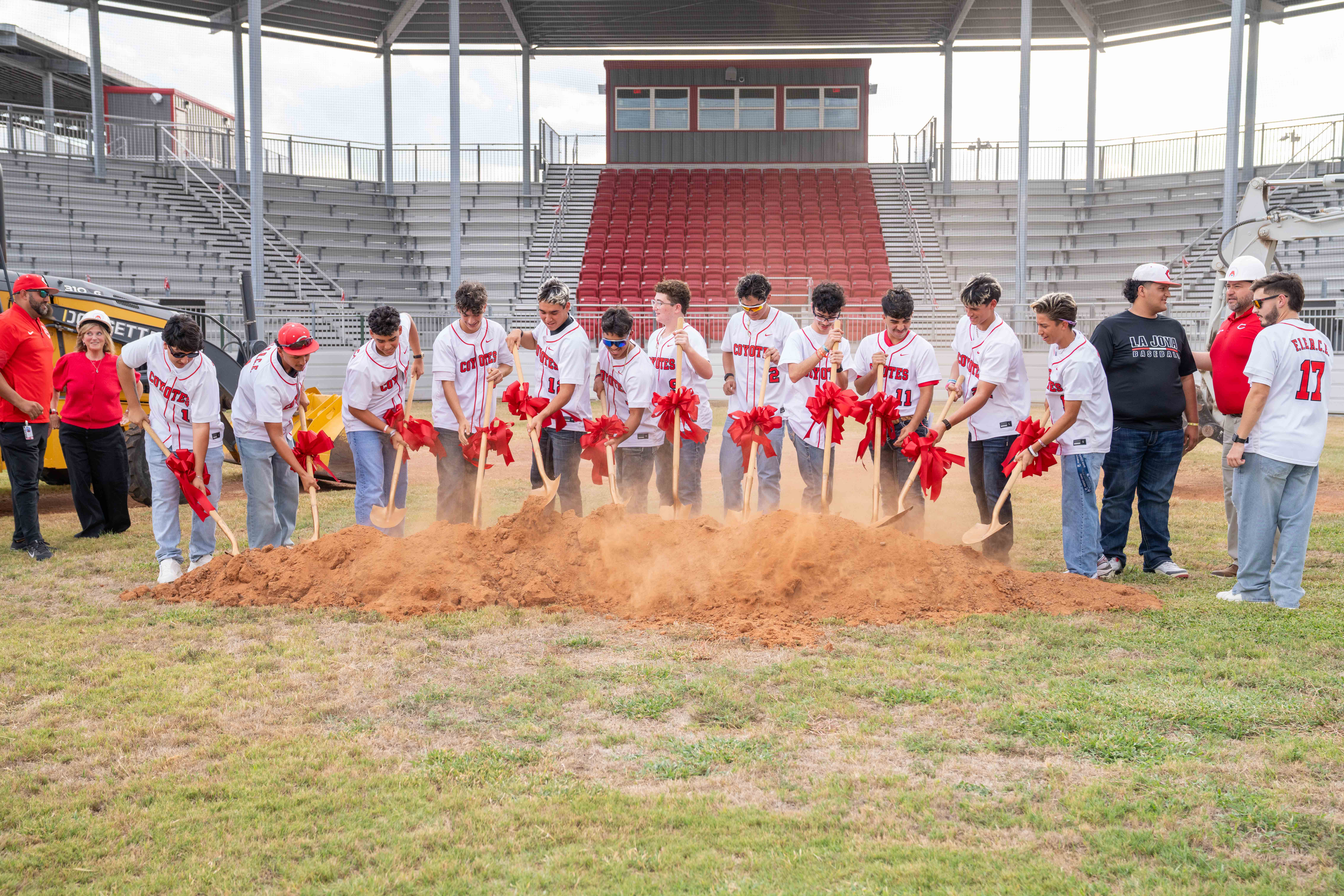 A baseball team performs a ceremonial groundbreaking in front of a stadium, with players digging into a pile of dirt using shovels adorned with red bows.