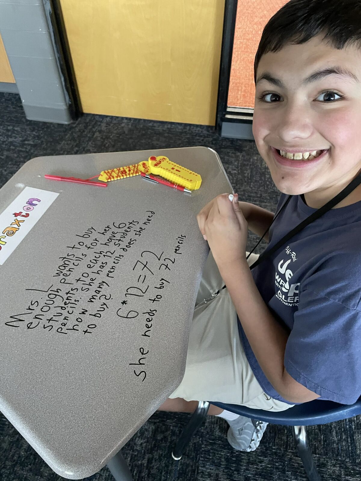 A smiling young boy sits at a school desk, proudly showing math work written in marker on the surface.