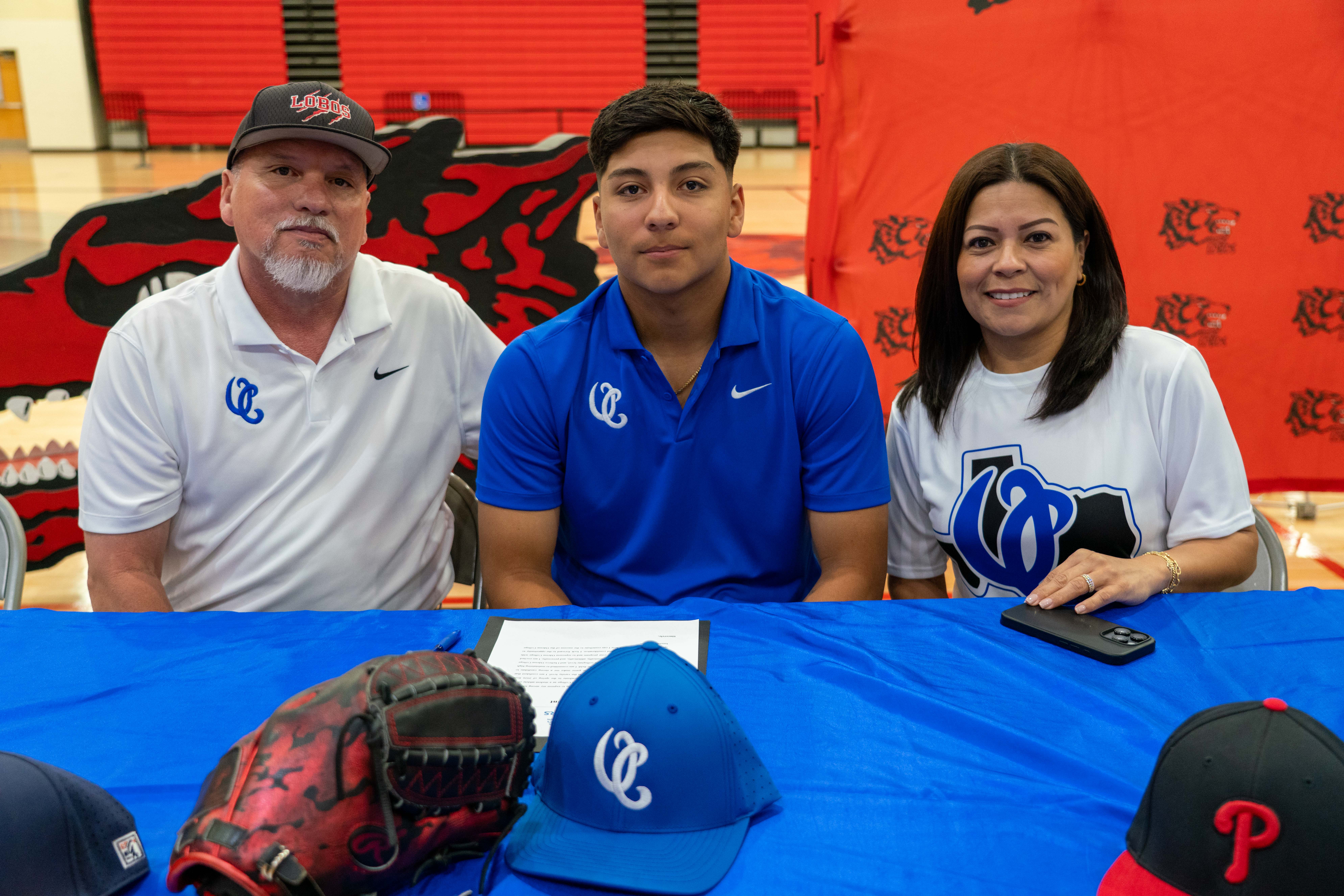 A young man in a blue polo shirt sits with his parents at a table, with baseball gear in the foreground.