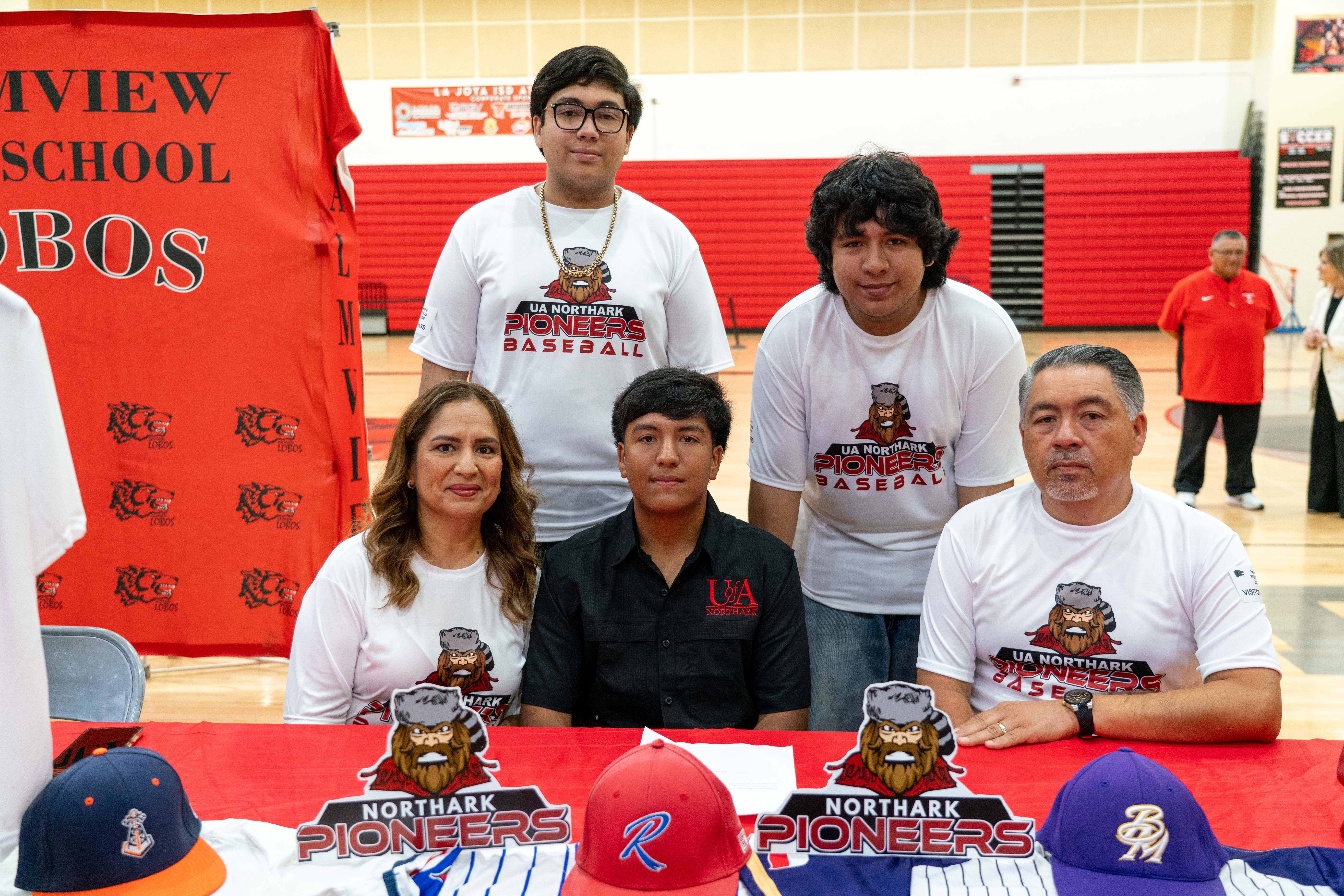A family poses for a photo at a table with baseball caps and jerseys, wearing shirts that say "UA Northark Pioneers Baseball."