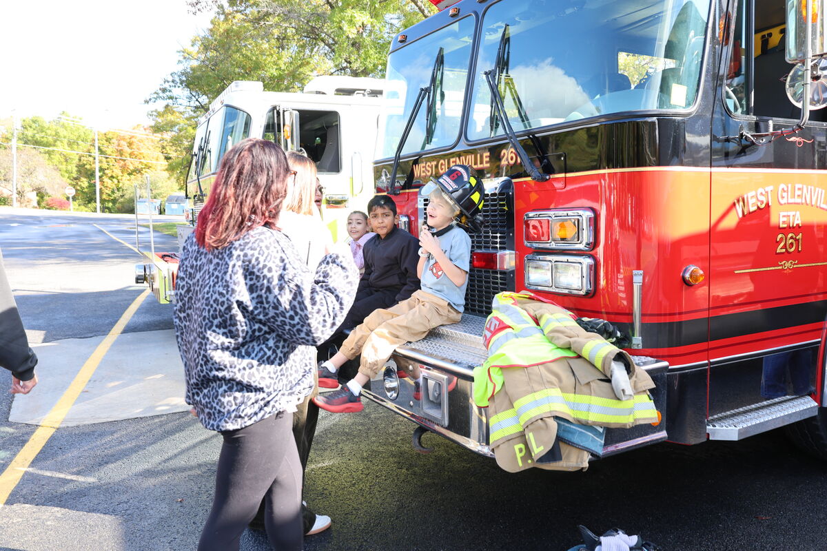 A child wearing a firefighter helmet sits on the front bumper of a red West Glenville fire truck, smiling as an adult talks to him.