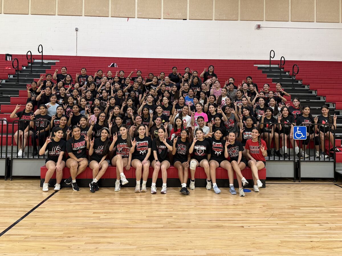 A large group of young women in athletic wear pose for a photo on bleachers in a gymnasium.
