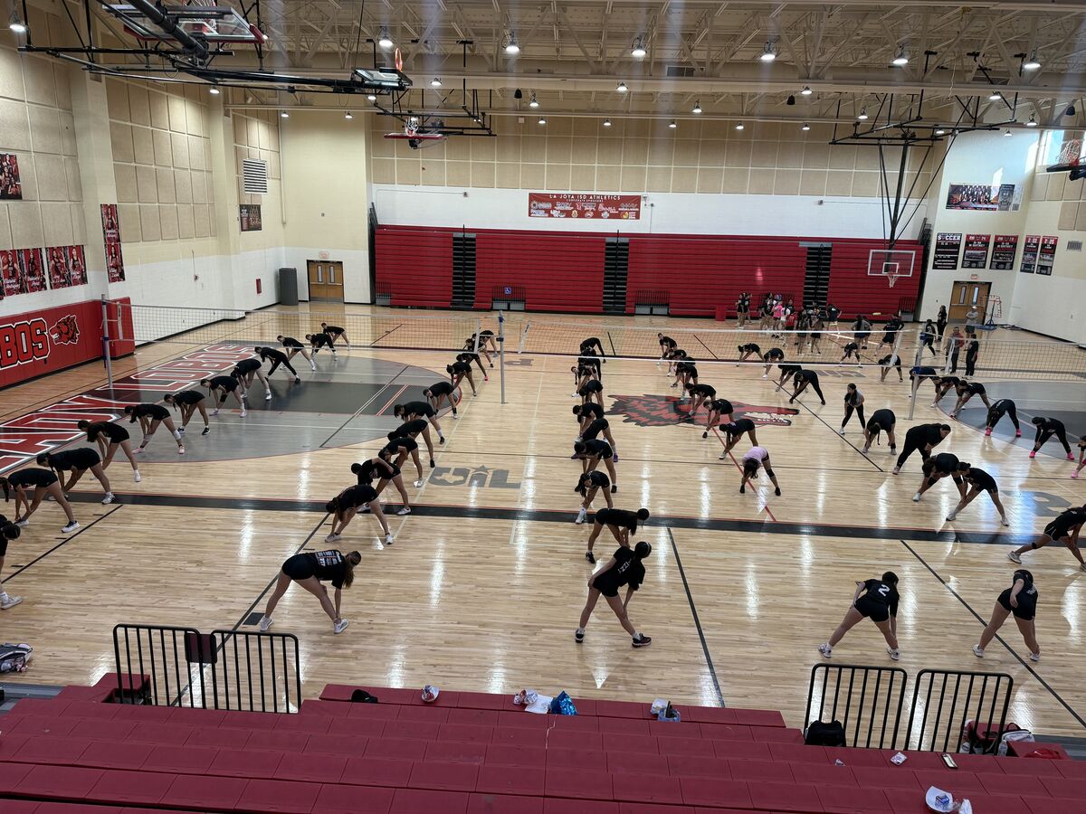 A group of athletes in black uniforms stretches on a wooden gymnasium floor.