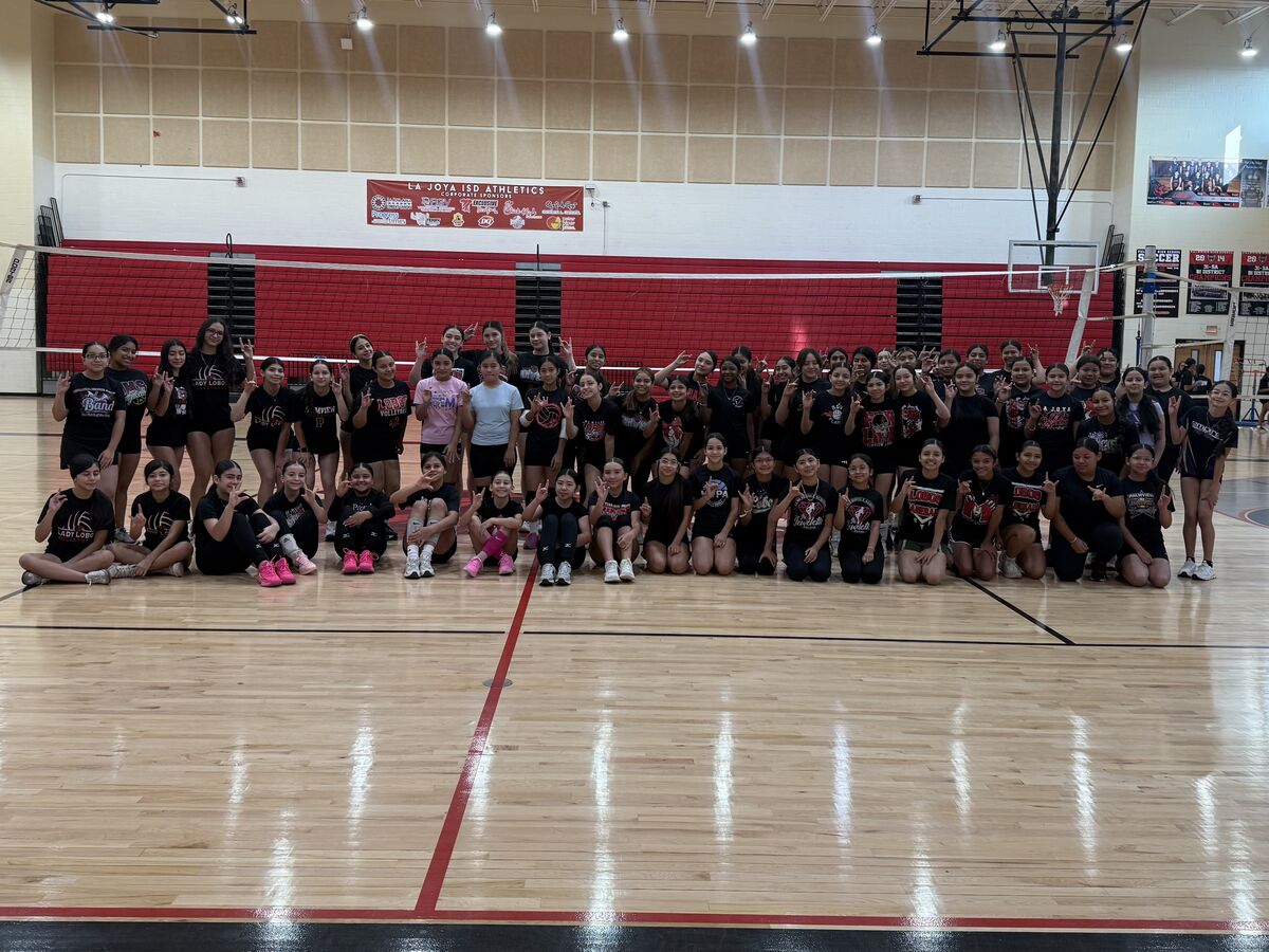 A large group of young women in black volleyball uniforms pose for a photo on a wooden gym floor.