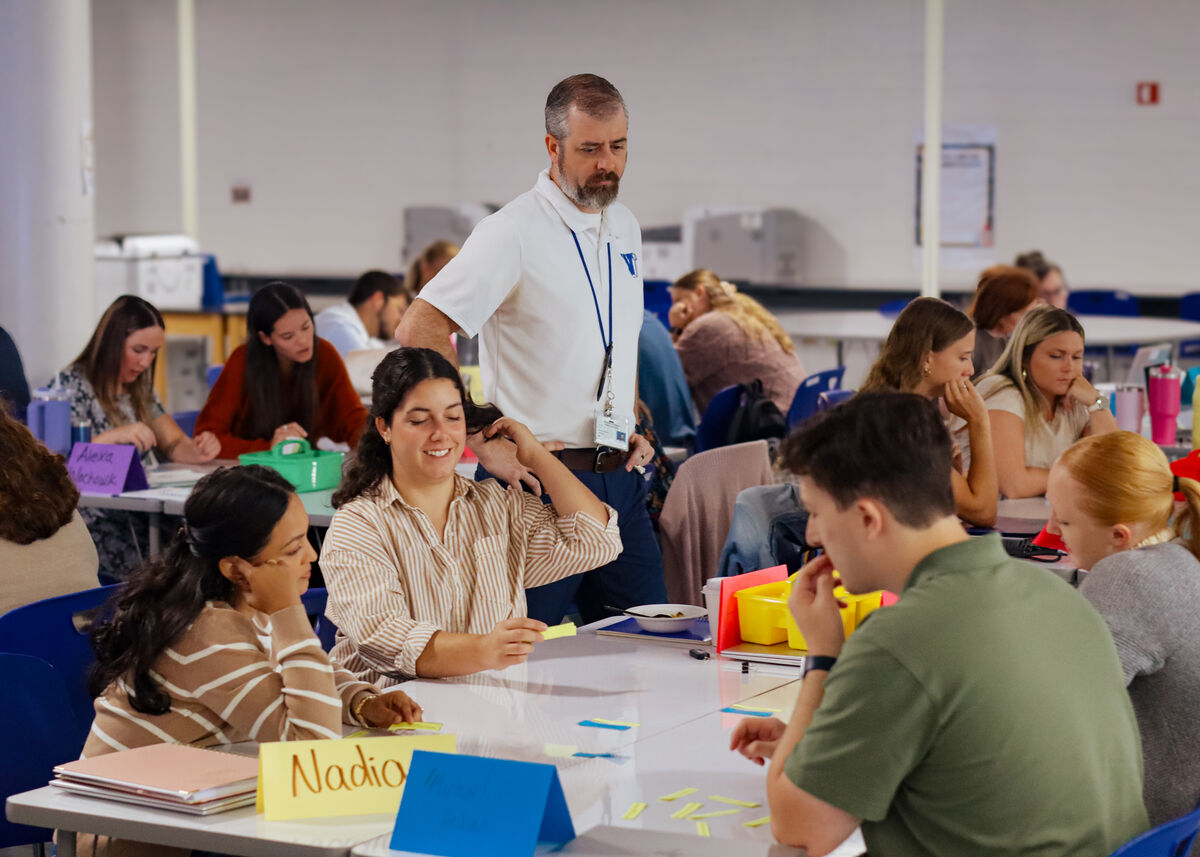 A man in a white polo shirt with a lanyard stands over a group of people seated at tables, observing their work.