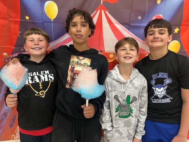 Four smiling boys stand together, each holding a fluffy ball of pink and blue cotton candy, in front of a circus-themed backdrop.