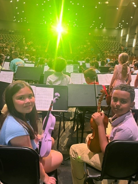 Two children hold violins, seated in an orchestra pit with sheet music.