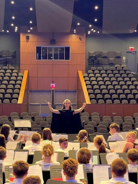 A conductor leads a band of young musicians in an auditorium with tiered seating.