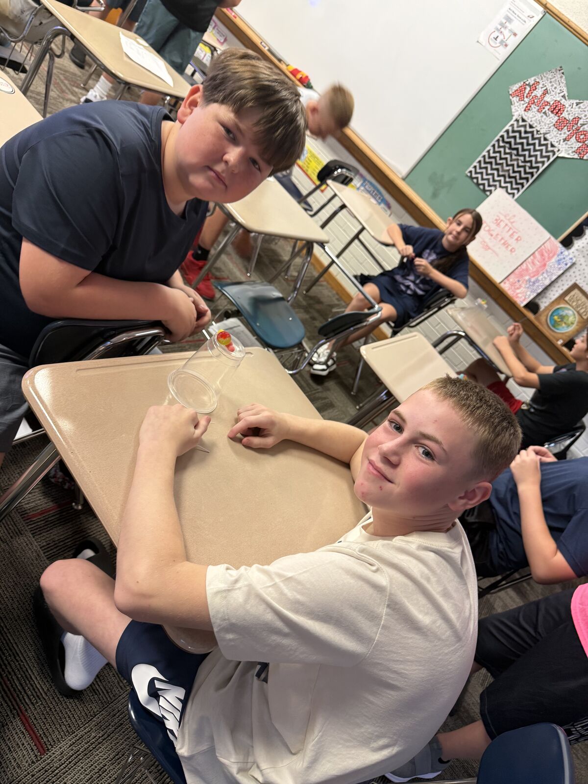 Two young boys are seated at desks in a classroom, looking towards the camera.