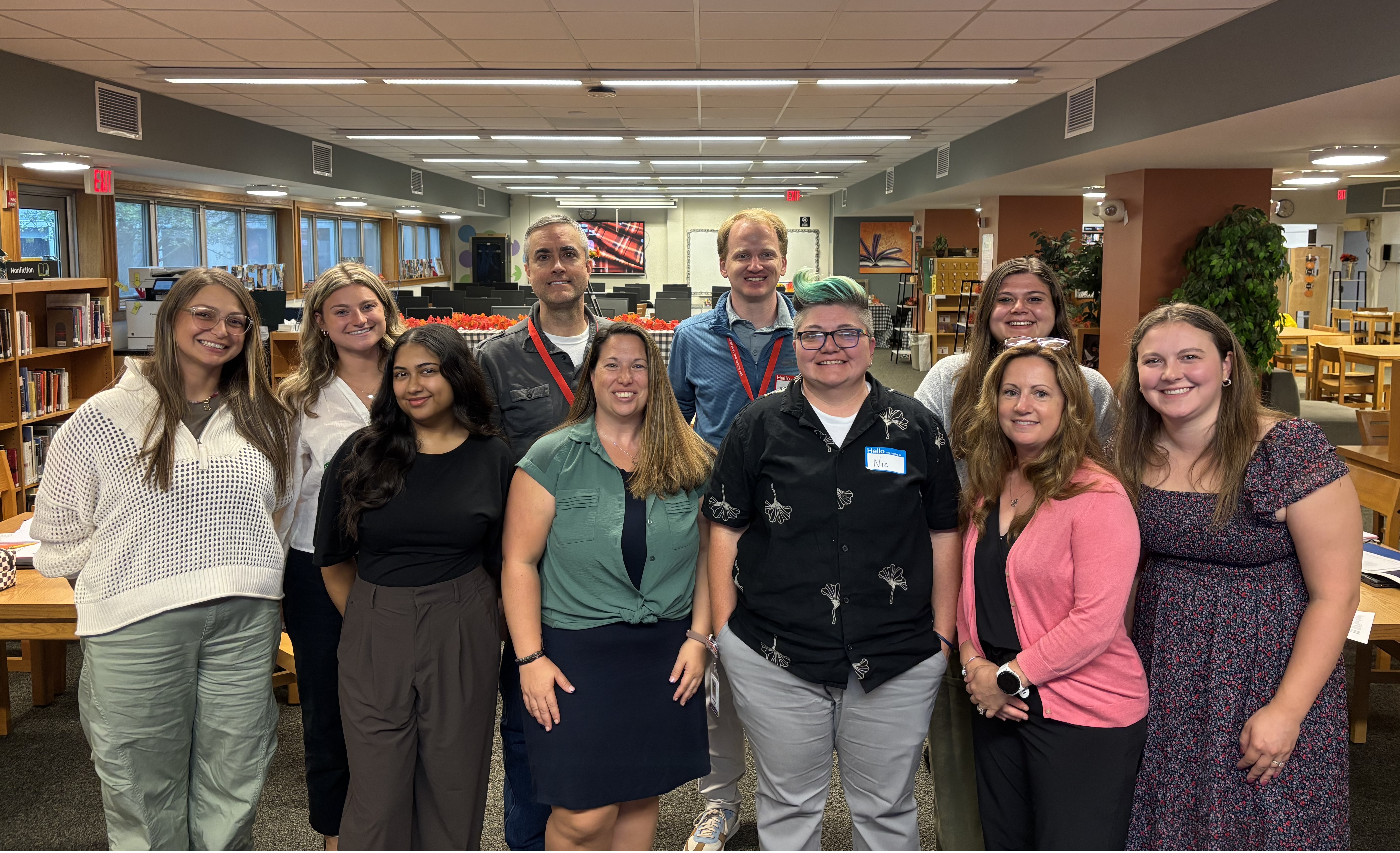 A group of ten diverse adults pose together smiling in a brightly lit school library.
