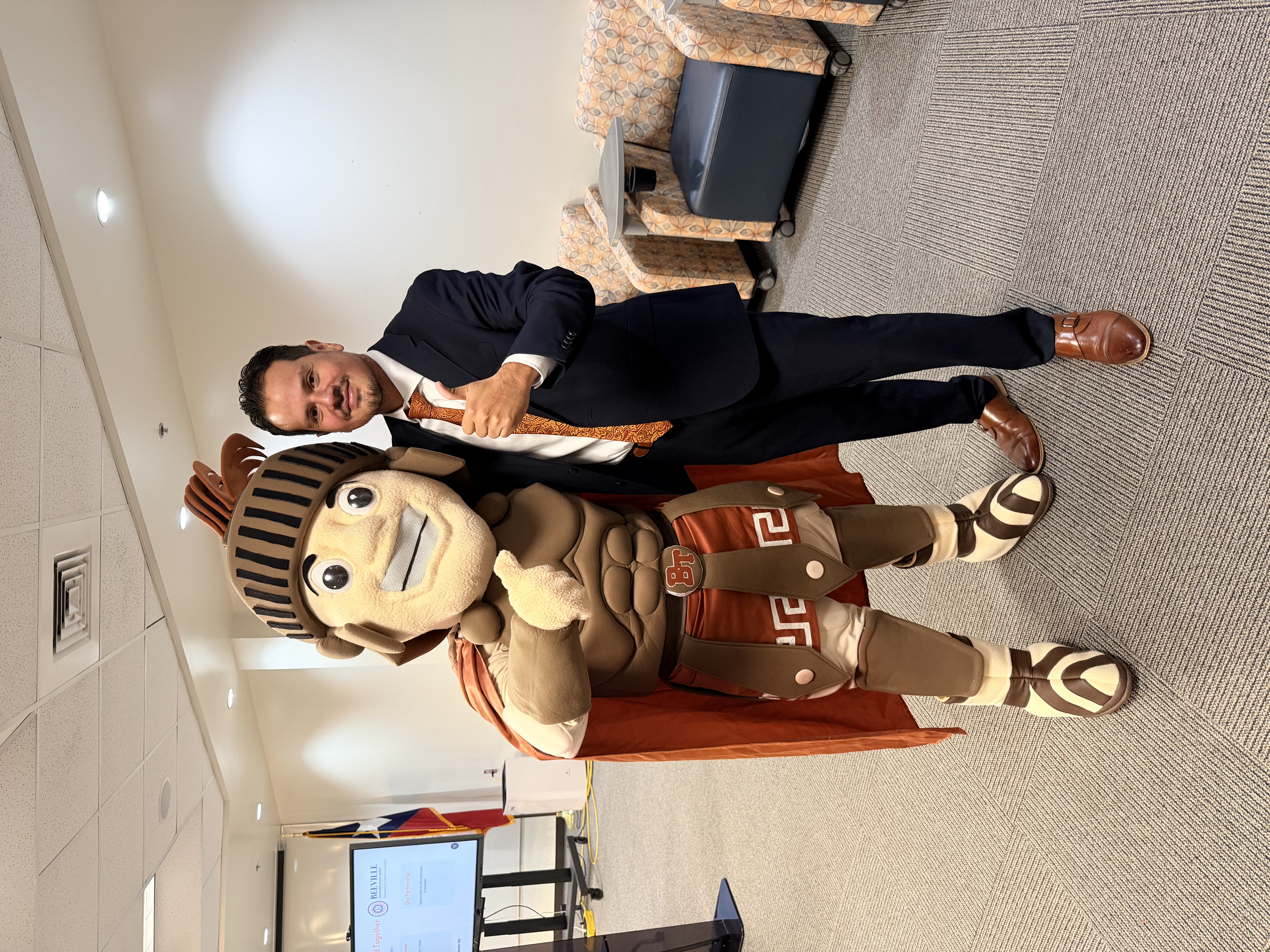 A man in a suit poses alongside a large mascot in a Trojan-style costume inside a building.