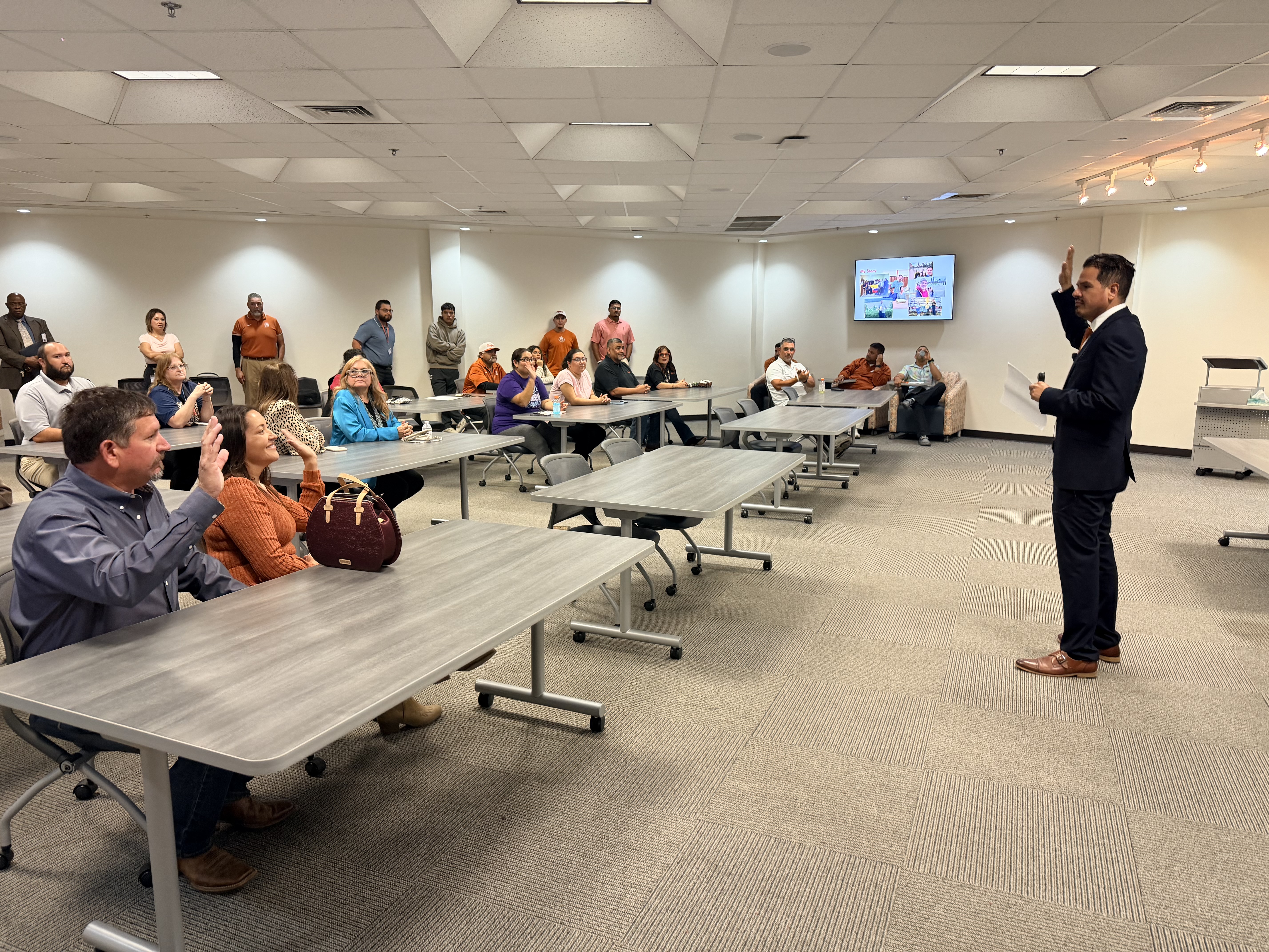 A speaker in a suit presents to an attentive audience seated at tables in a brightly lit conference room.