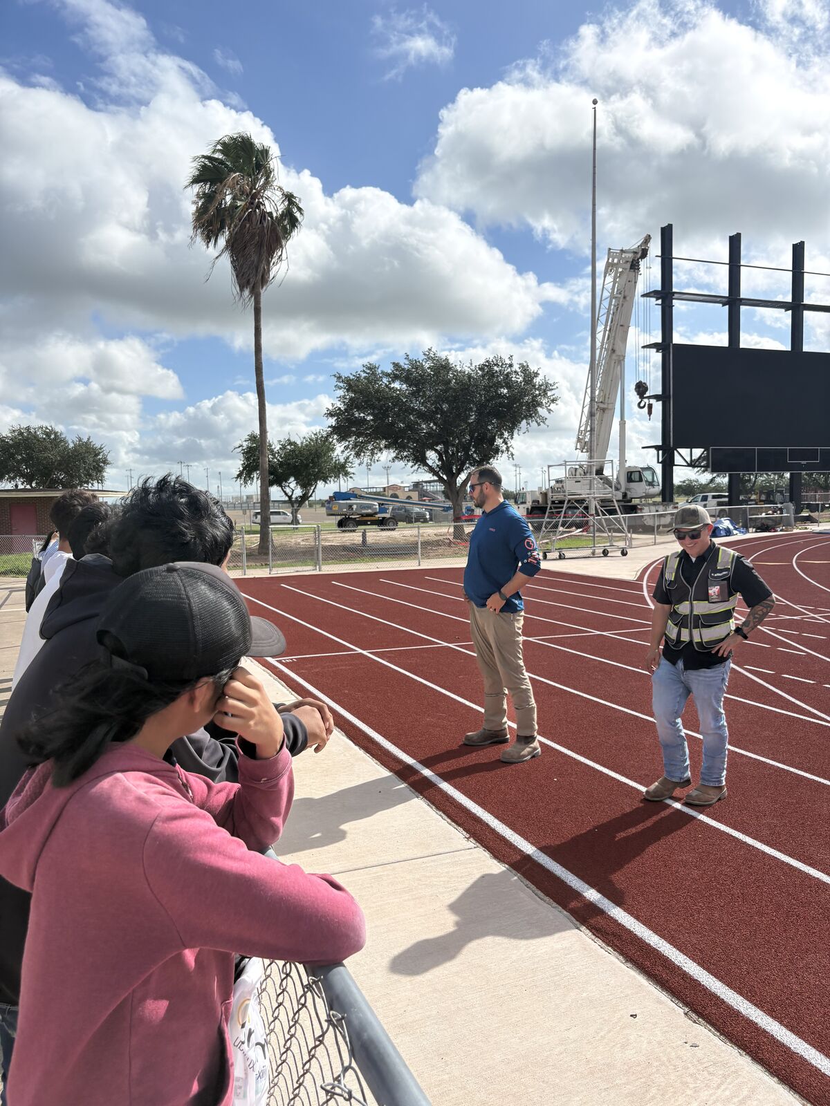 People stand on a red running track with white lines, under a cloudy blue sky.