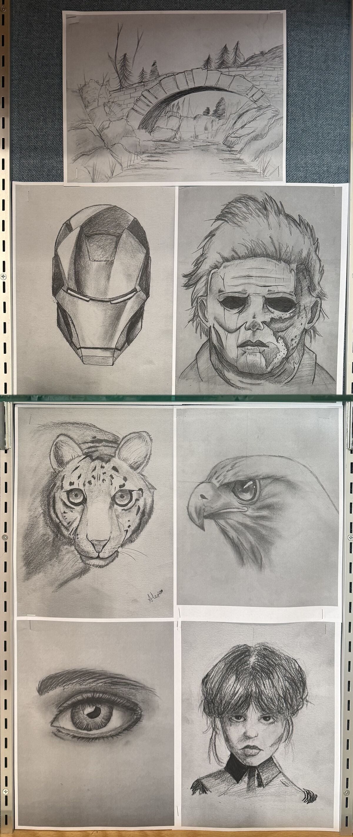 A collection of six pencil drawings pinned to a bulletin board, featuring a bridge, an Iron Man mask, a horror movie mask, a snow leopard, an eagle, an eye, and a portrait of a person.