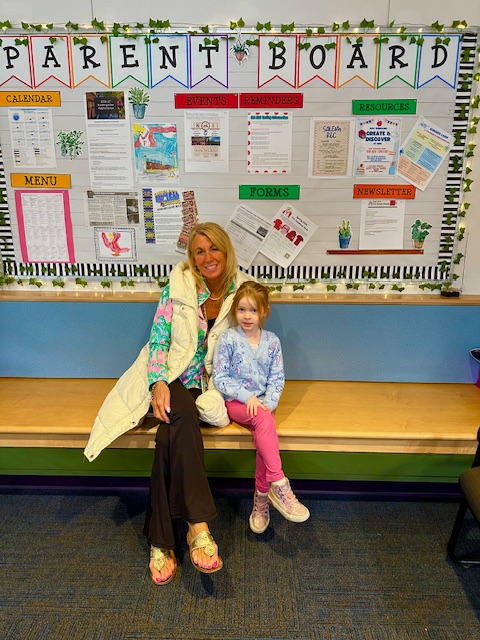 A woman and a young girl sit on a wooden bench in front of a "PARENT BOARD" with various notices.
