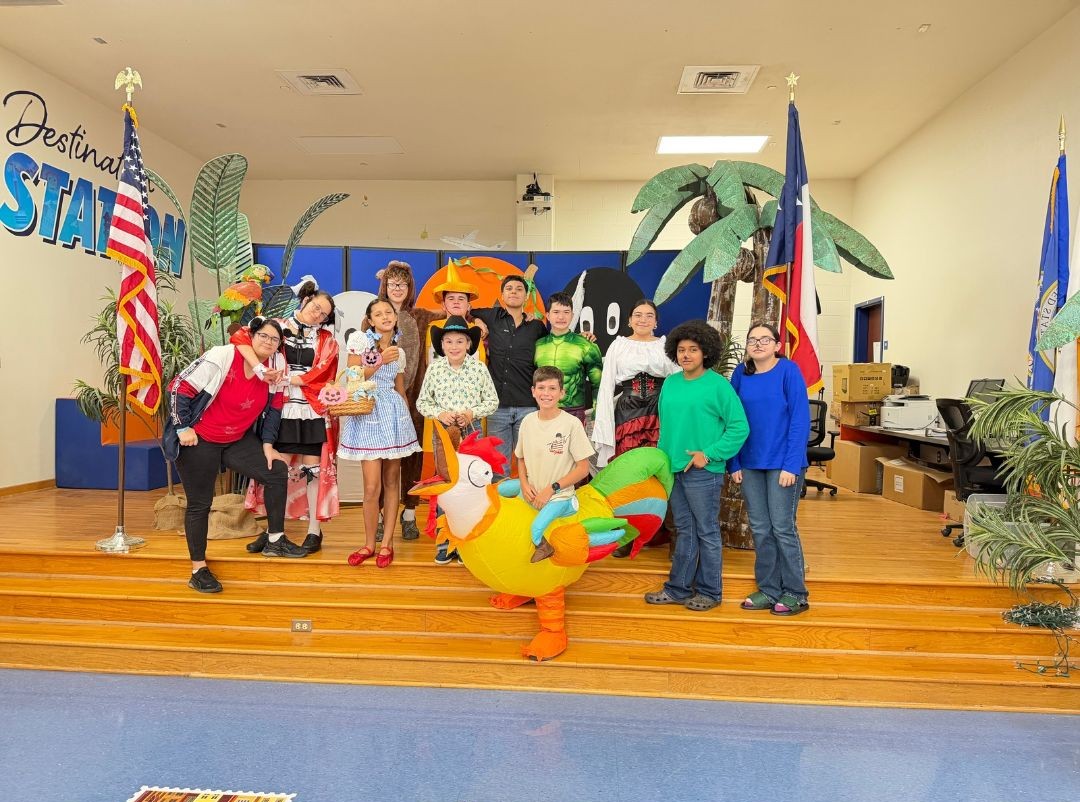 A group of students poses on a stage in creative costumes, with American and Texas flags on the sides.