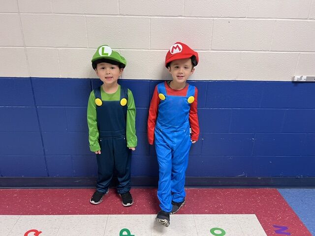 Two young children stand side by side against a cinder block wall, dressed in Mario and Luigi costumes.