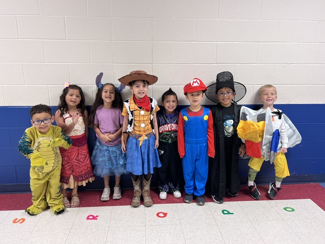 Seven children in diverse costumes stand in a line against a white and blue brick wall.