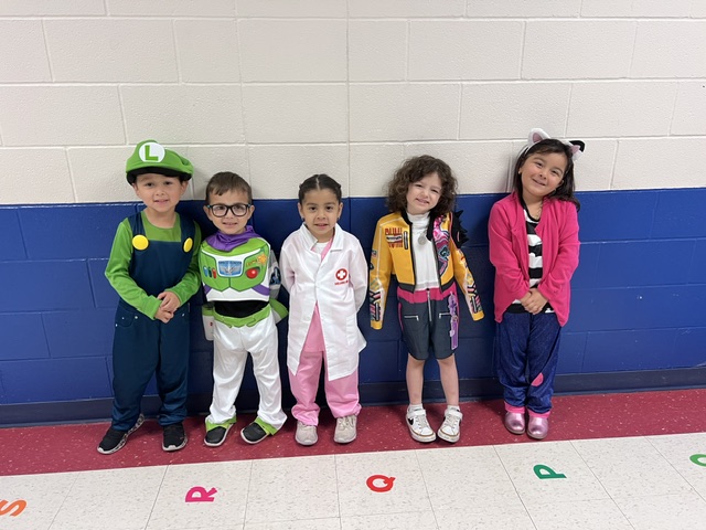 Five children pose happily in a hallway, dressed in various colorful costumes including Luigi, Buzz Lightyear, and a doctor.