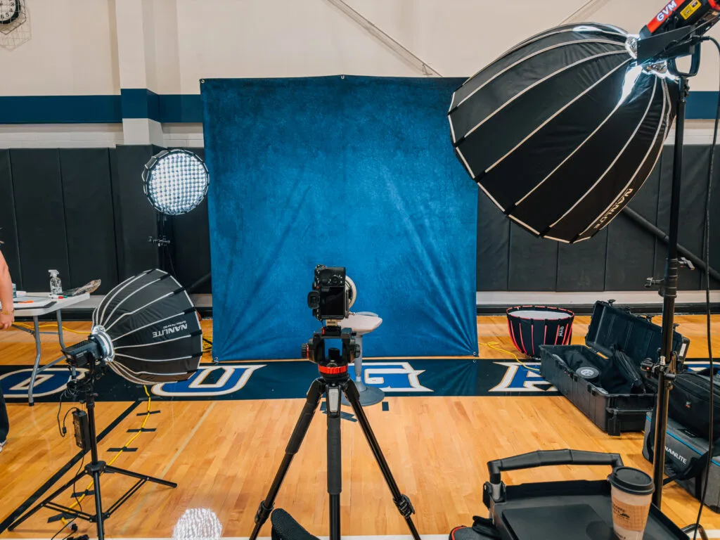 A photography setup in a gymnasium with a blue backdrop, camera on a tripod, and studio lights.