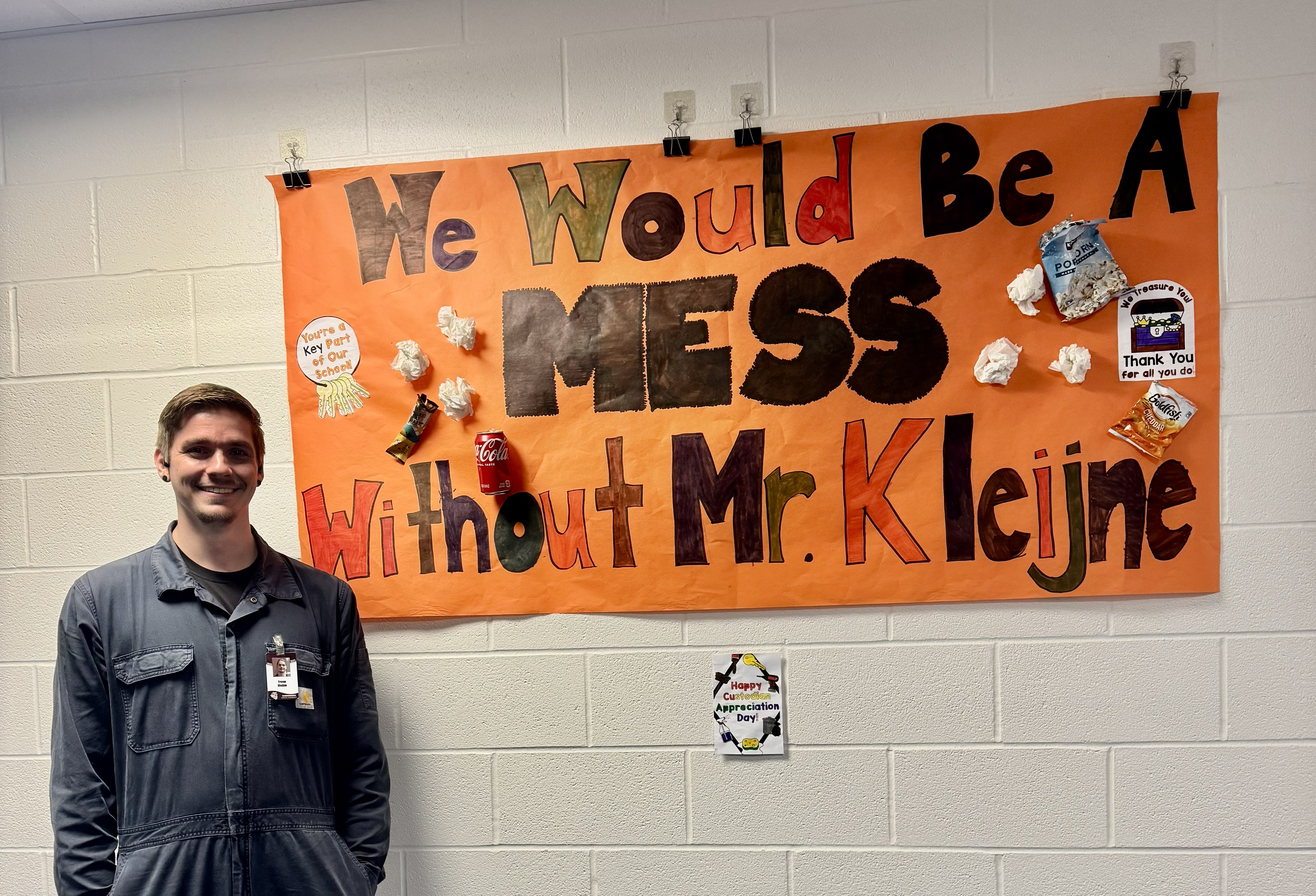A smiling man stands in front of an orange banner that reads 'We Would Be A MESS Without Mr. Kleijne'.